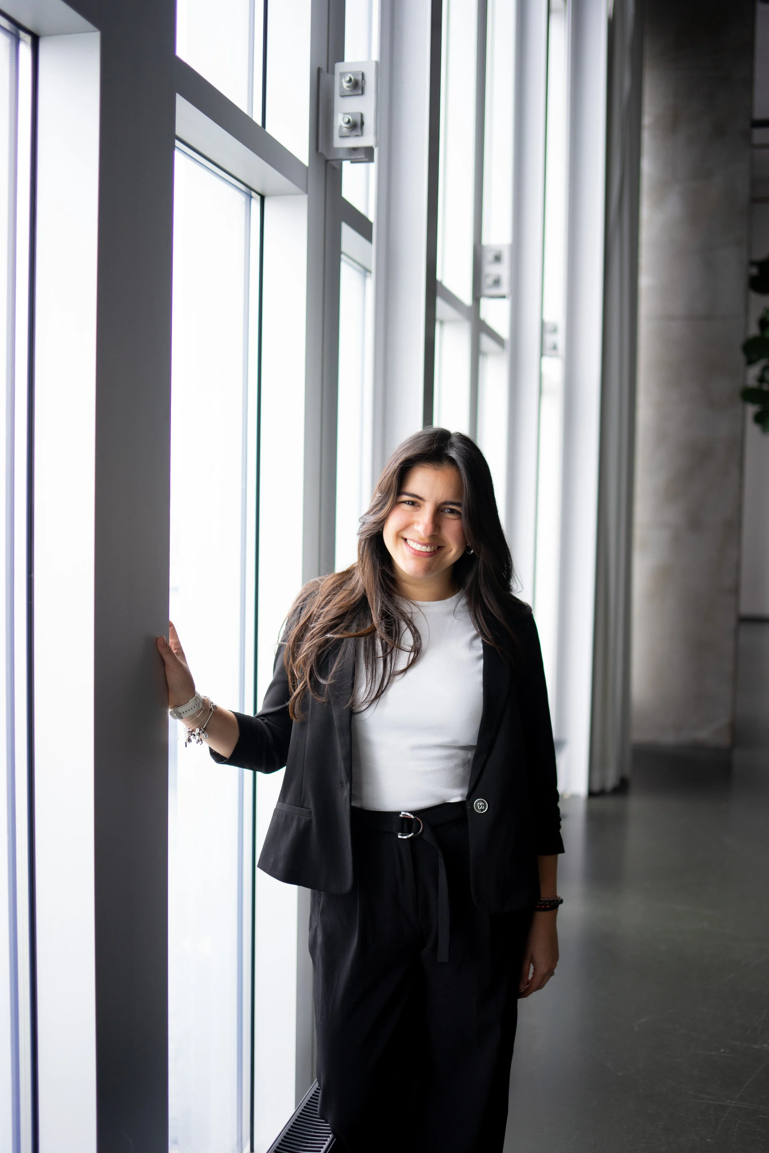 A businesswoman smiling and standing in front of a row of large glass windows, wearing a black blazer, white shirt, and black pants with a belt, inside a modern building.