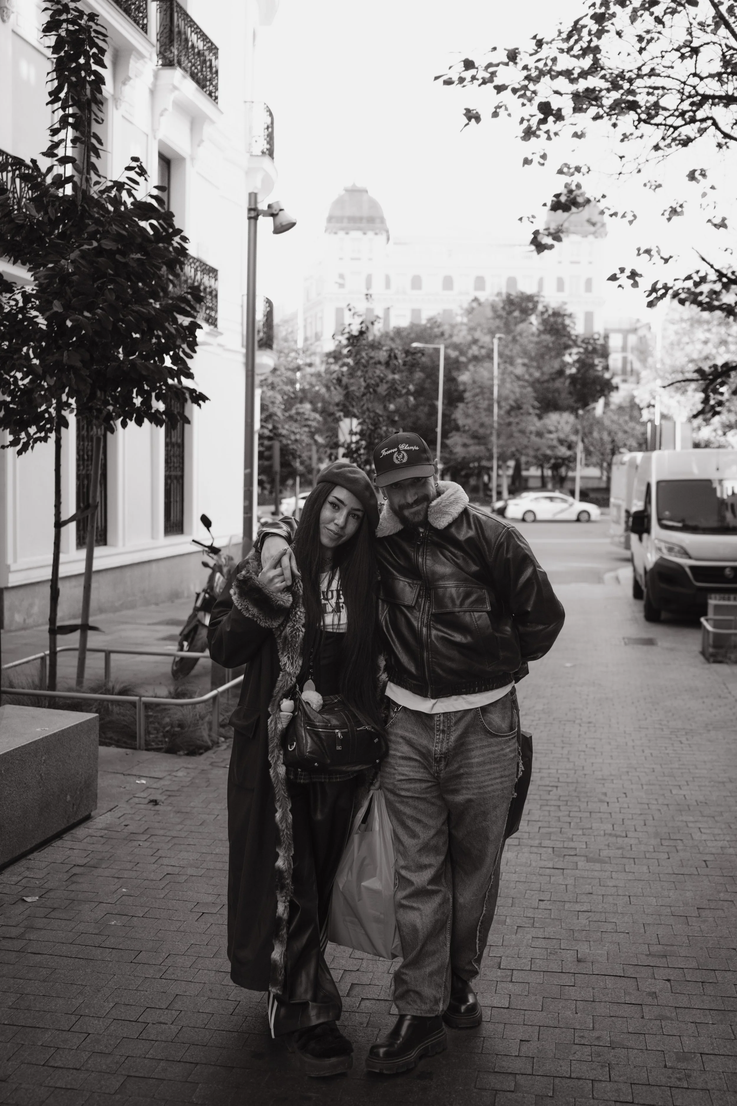 A black-and-white photo of a young woman and a man walking together on a city sidewalk, smiling and embracing each other. The woman has long hair and is wearing a fur-trimmed jacket and beanie, while the man has a beard, is wearing a cap, leather jac