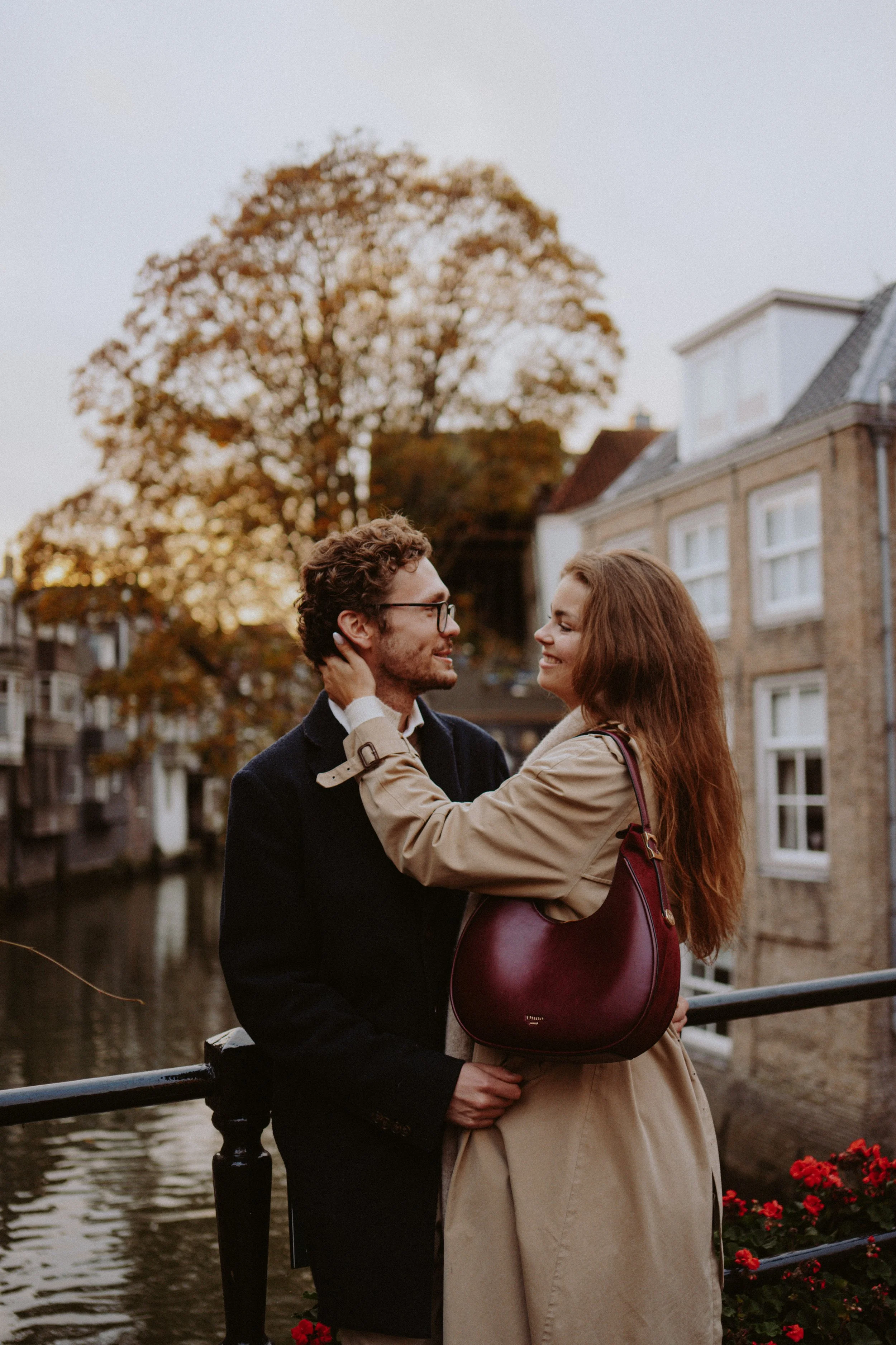 A man and woman happily looking at each other on a bridge with an autumn tree in the background.