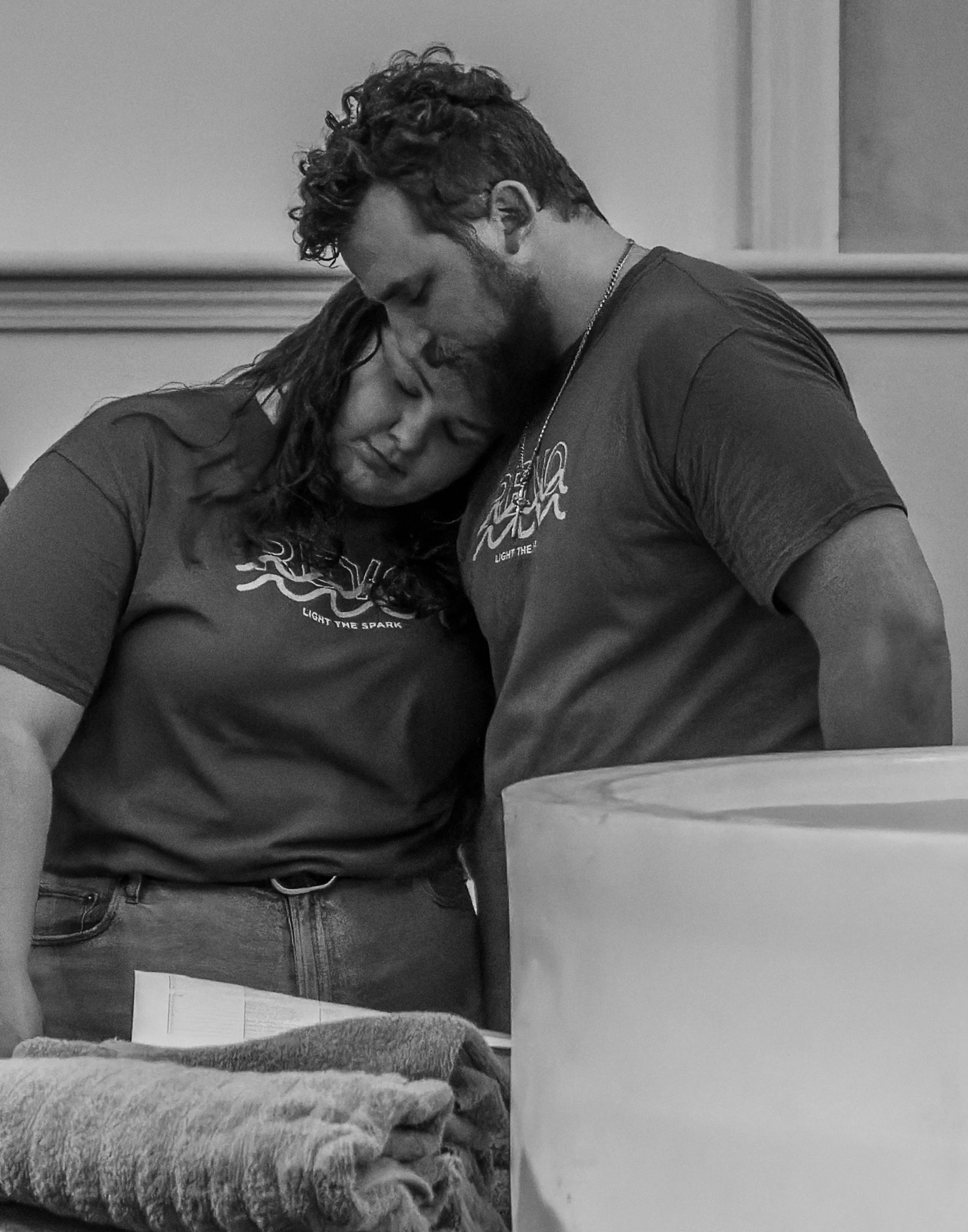 A man and woman are closely leaning their foreheads together, with eyes closed, in a moment of intimacy or comfort. They are indoors, with the woman in a long-sleeved shirt and the man in a t-shirt. The background shows a wall with molding.