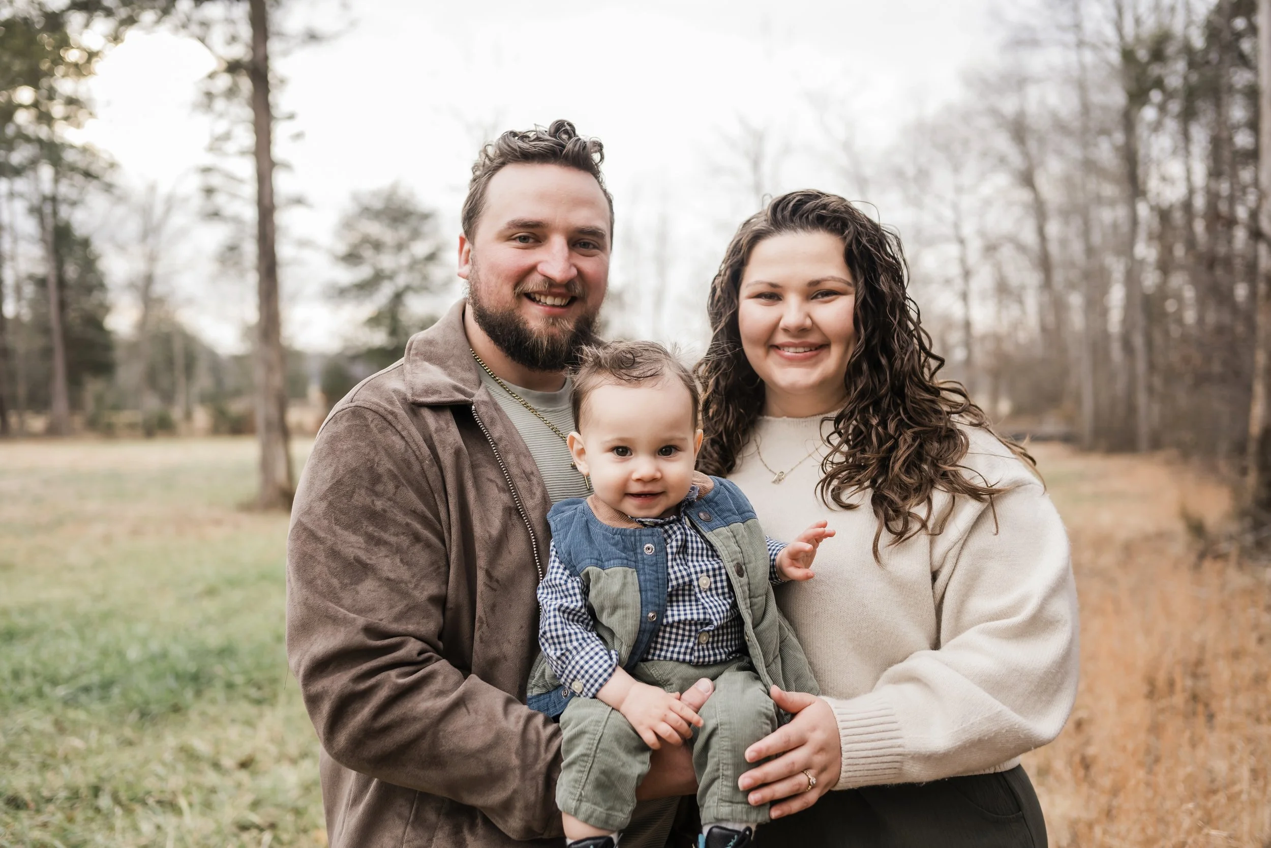 A happy family of three standing outdoors in a wooded area during fall. The father has a beard and is smiling at the camera. The mother has curly hair and is smiling as well. The young child, who is being held by both parents, has curly hair and is looking at the camera with a curious expression.