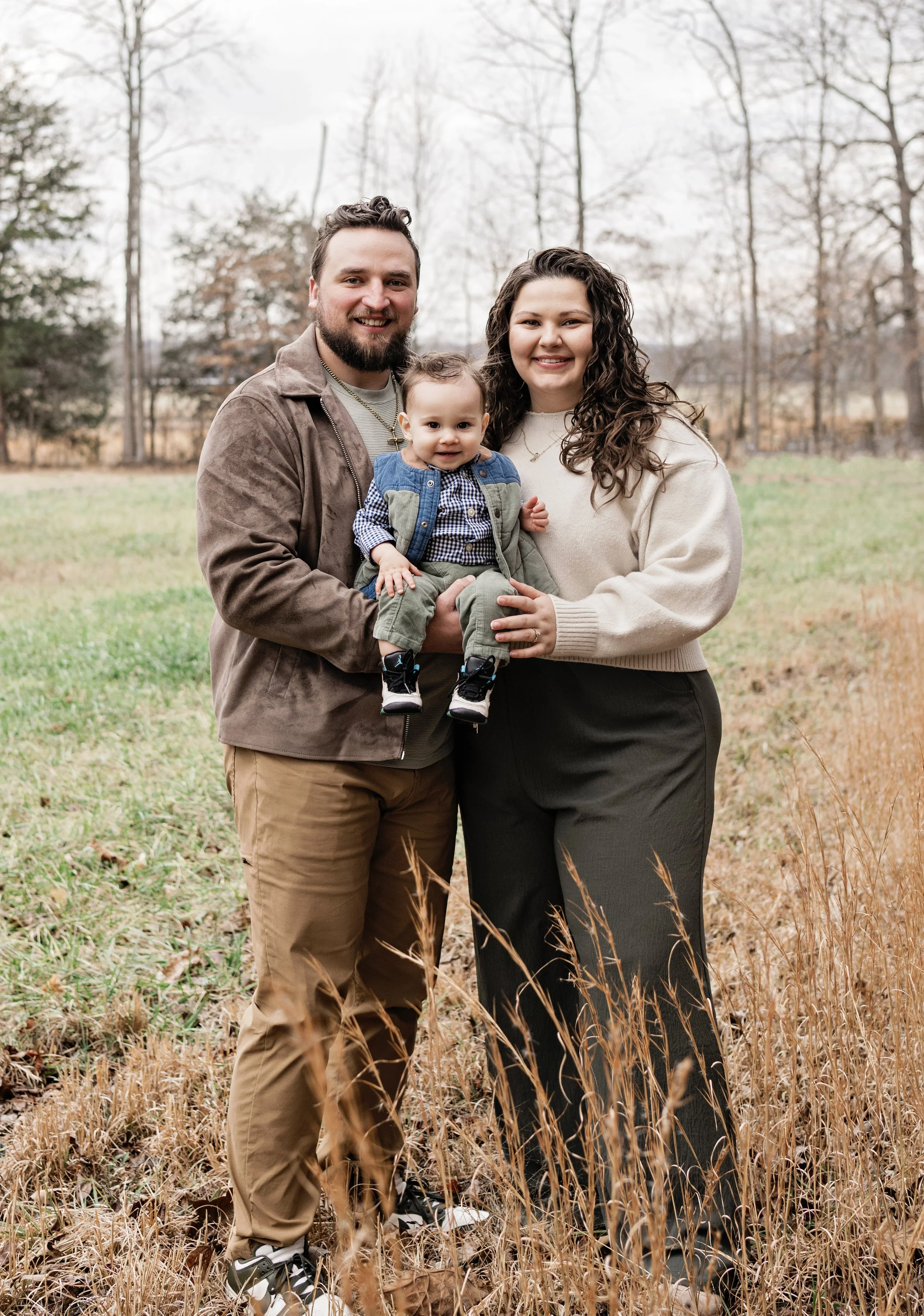 A family of three standing outdoors in a field with tall grass and trees in the background. The man has a beard and is wearing a brown jacket, the woman has curly hair and is wearing a cream sweater, and the toddler is sitting on the man's arm, wearing a checkered shirt, a vest, and sneakers.