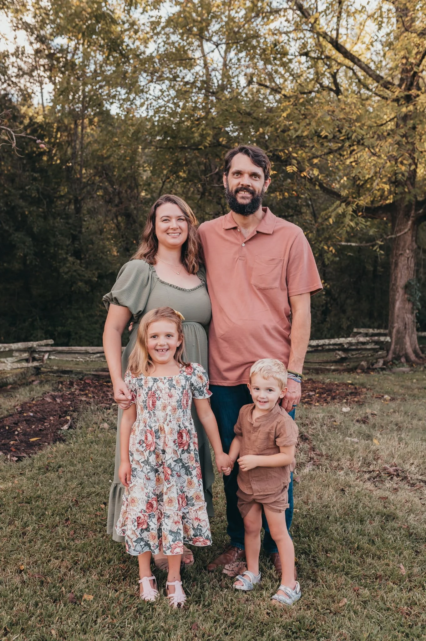 A family photo taken outdoors during daytime, featuring two adults and three children standing on a grassy area with trees in the background.