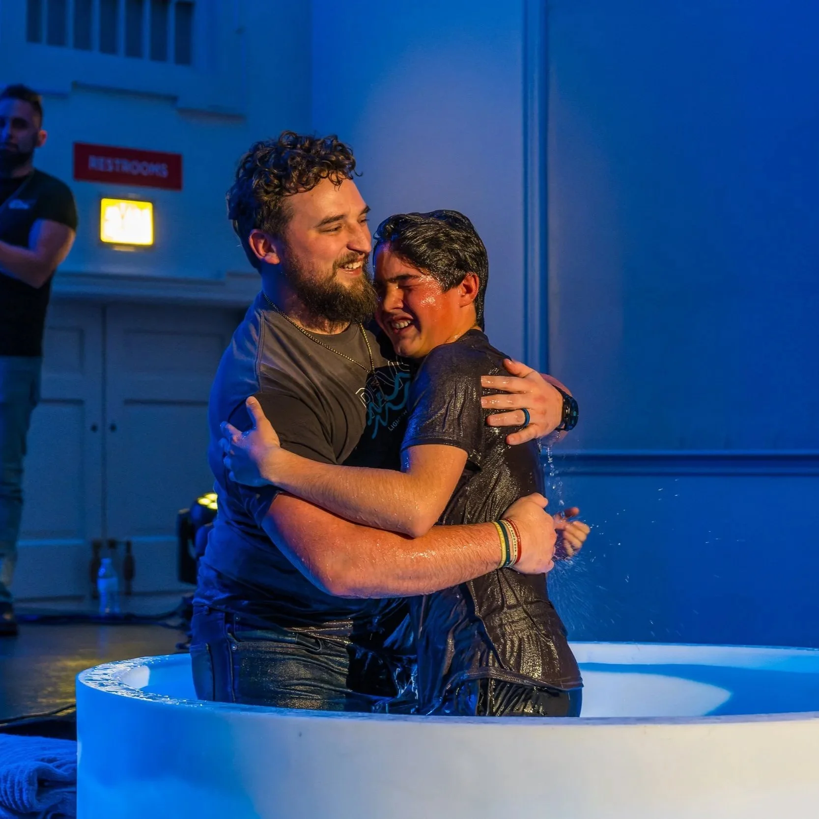 Two men embracing in a baptismal pool, smiling and joyful, during a baptism ceremony indoors with a blue-lit background.