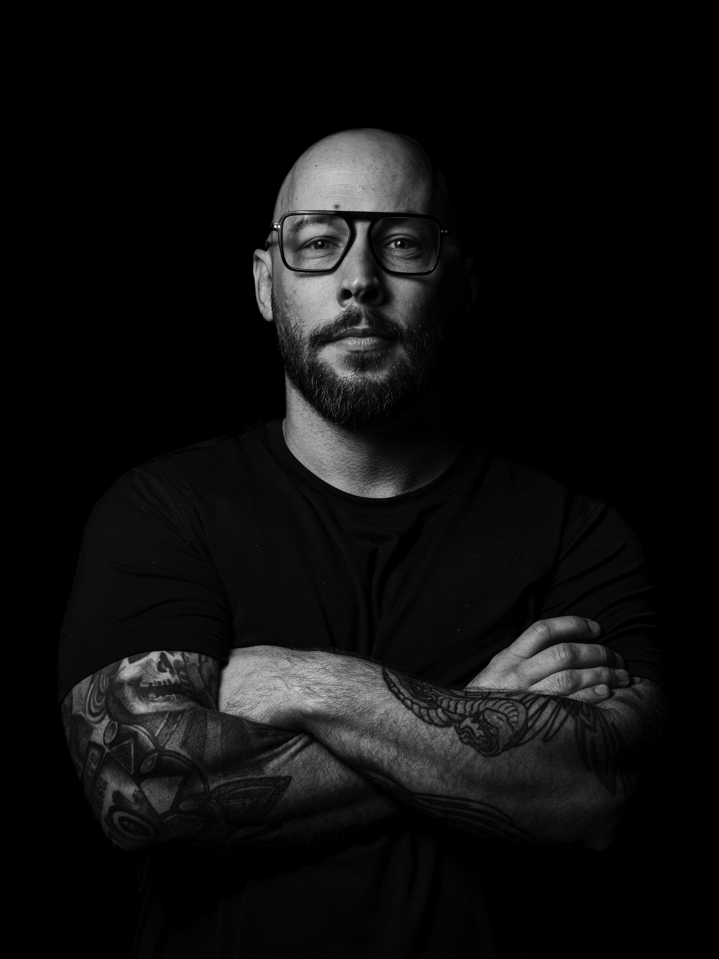 Black and white photo of a man with a beard and tattoos, wearing glasses, crossing his arms, against a dark background.