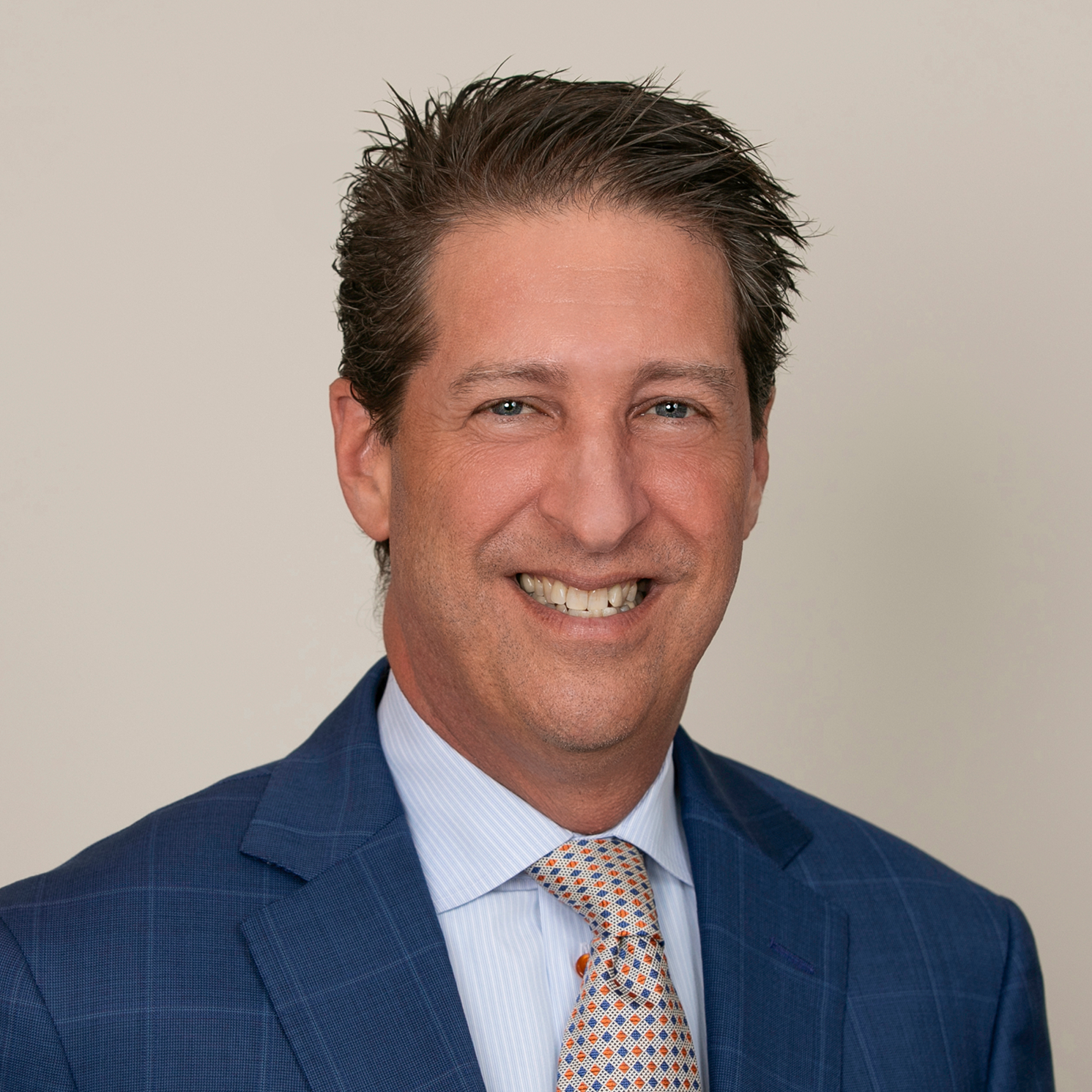 A middle-aged man in a blue checkered suit, light blue shirt, and patterned tie, smiling with blue eyes and brown hair styled back, against a plain white background.