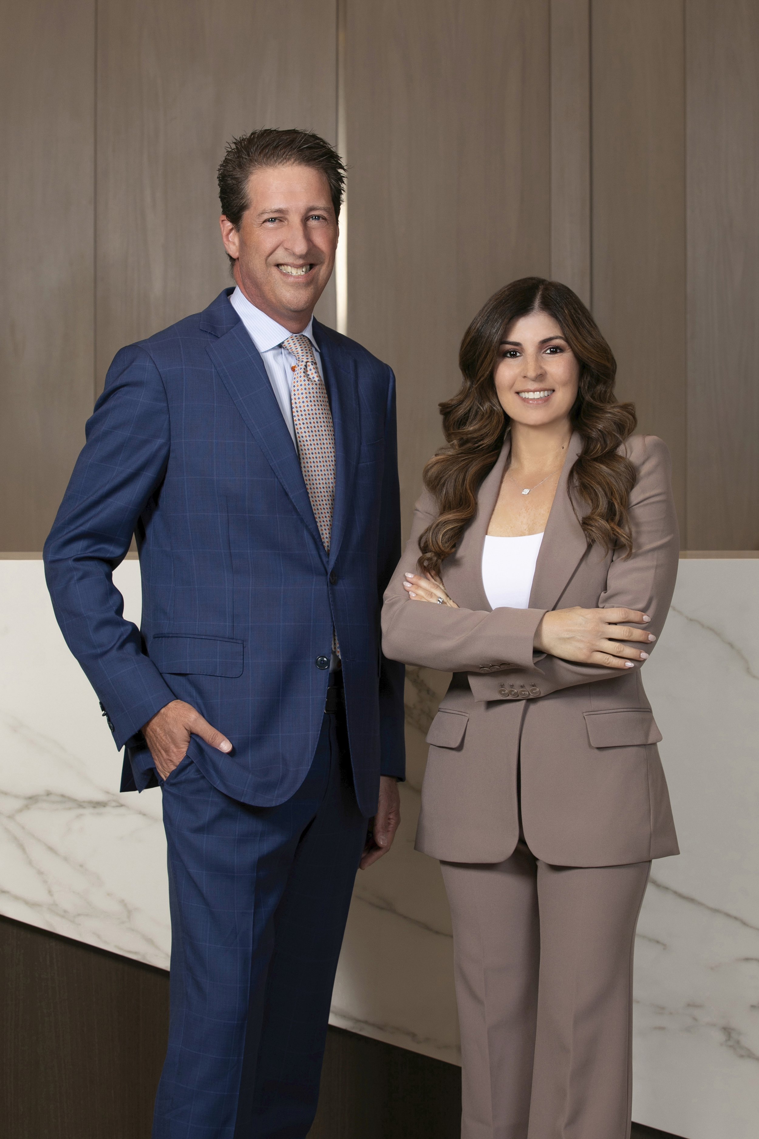 A man in a blue checkered suit and a woman in a brown suit standing side by side in an office setting, smiling at the camera.