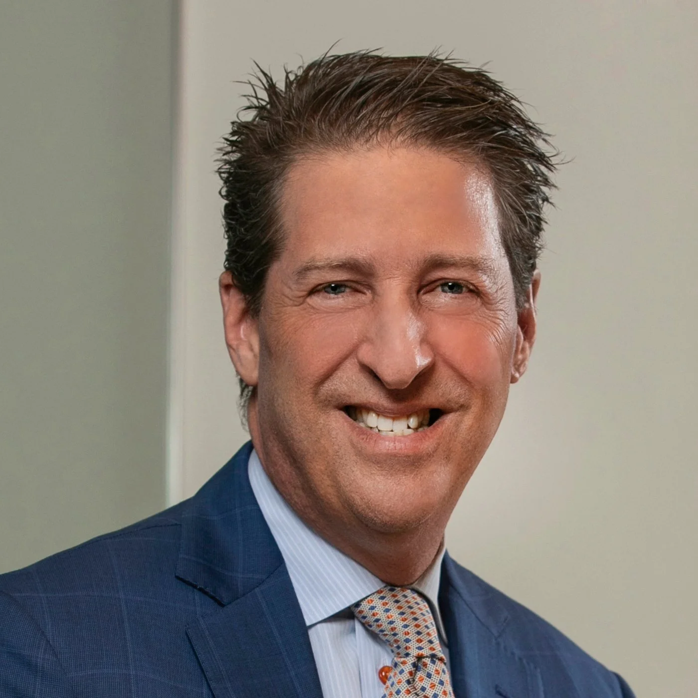 A smiling man with brown hair styled upwards, wearing a blue suit, a light blue shirt, and a patterned tie, against a background with neutral colors.