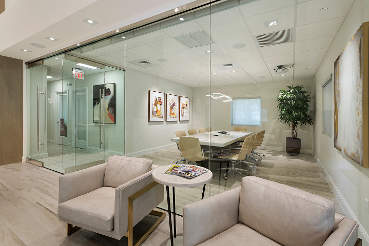 Modern conference room with a glass wall, beige chairs around a large table, artwork on the walls, a window with blinds, and a potted plant.