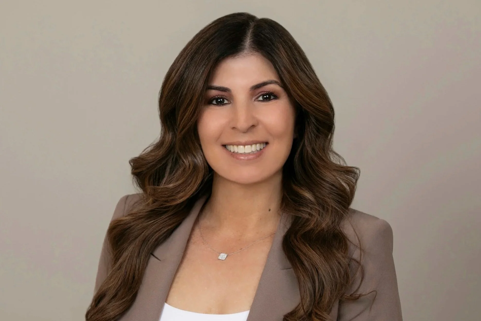 A woman with long wavy brown hair smiling, wearing a taupe blazer and a locket necklace, against a plain background.