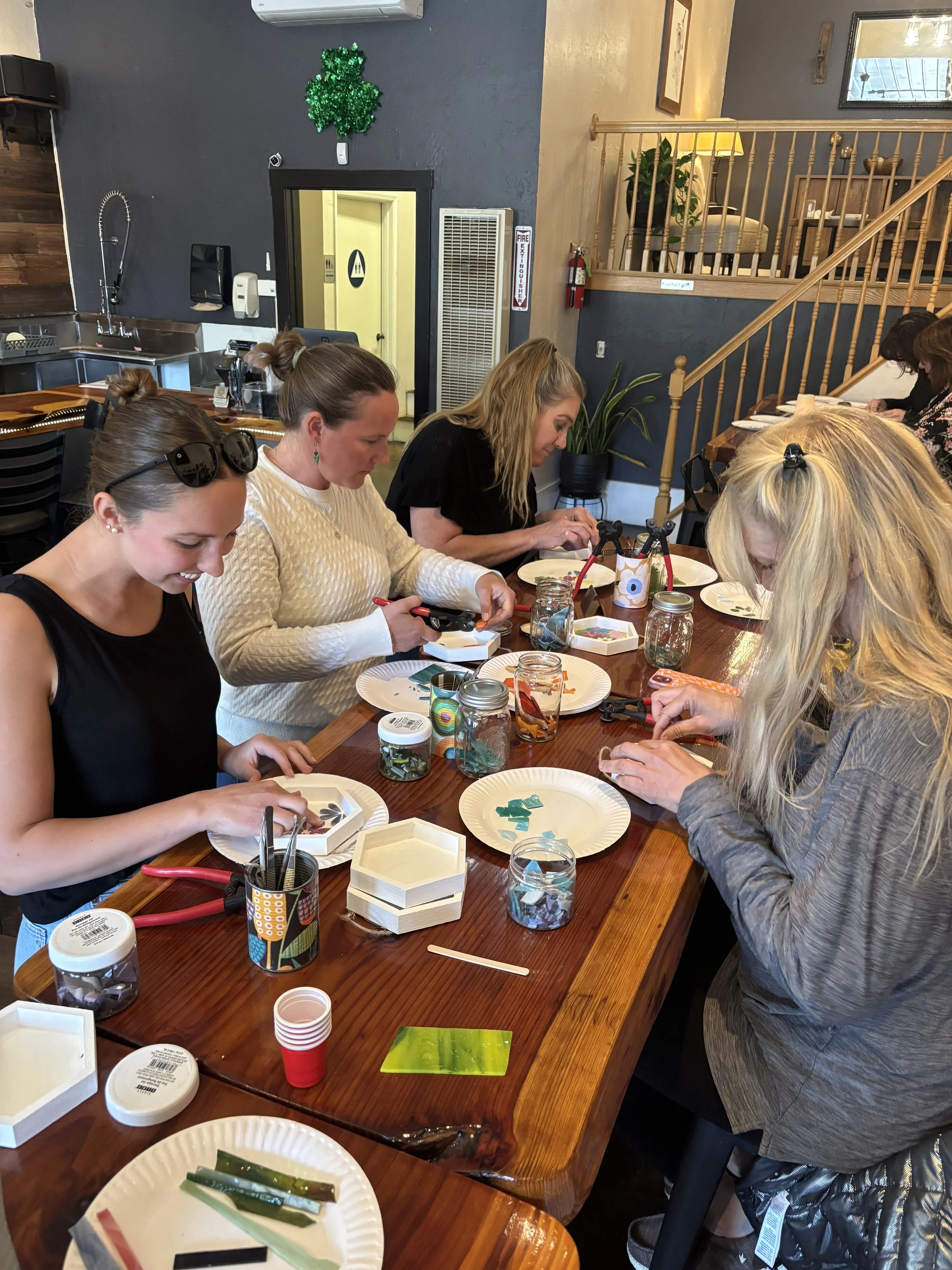 Group of women sitting around a wooden table engaged in a craft activity with glass pieces, glue, and tools; casual indoor setting with stairs and plants in the background.