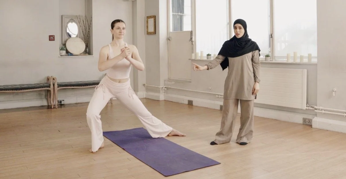Two women practicing martial arts indoors, one in a yoga pose and the other giving instructions, in a bright room with windows and minimal decor.