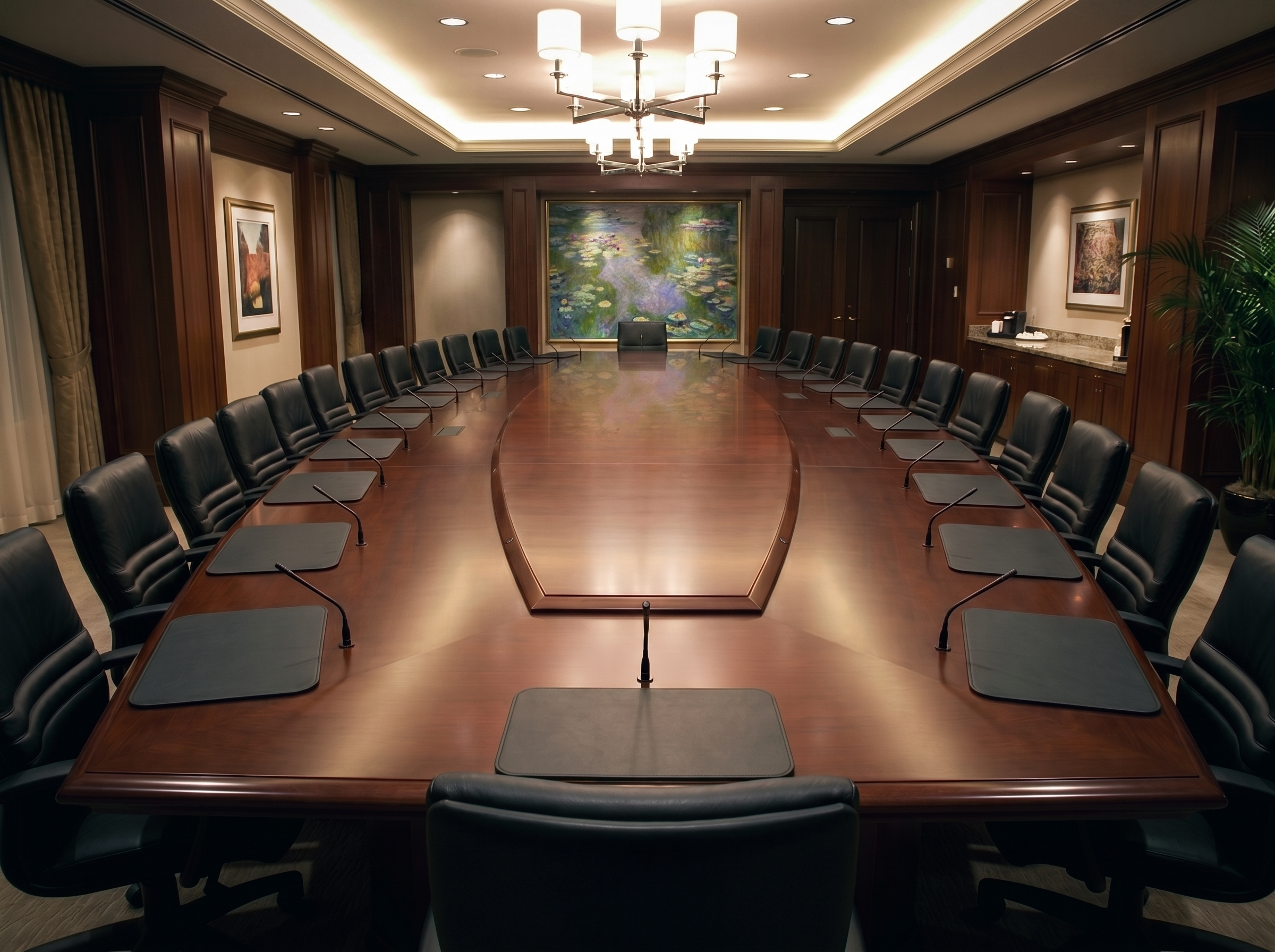 A large, oval-shaped conference room with a polished wooden table surrounded by black leather chairs. The room features wood-paneled walls, modern artwork, and a ceiling with recessed lighting and a chandelier.