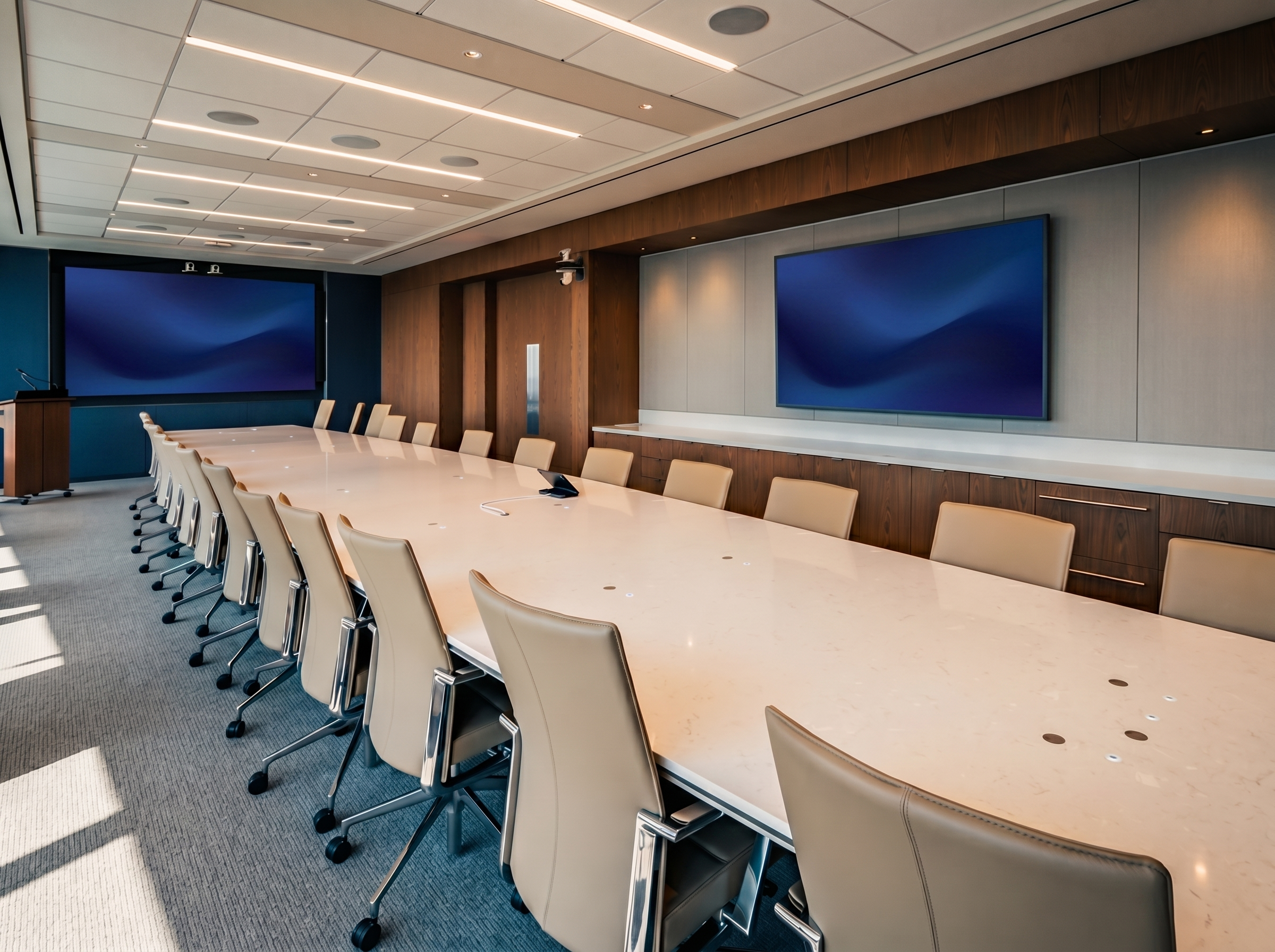A modern conference room with brick and acoustic panel walls, a large digital display, multiple desks and chairs arranged for a meeting or presentation.