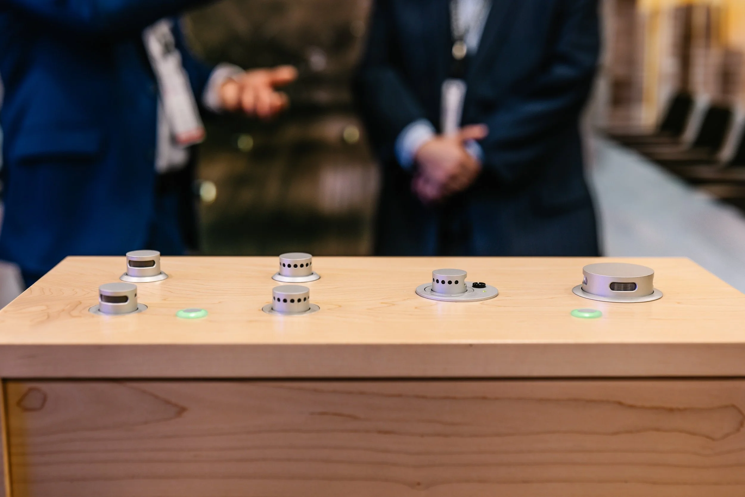 Blurry image of two people in suits standing behind a wooden table with modern game controllers or devices on it.