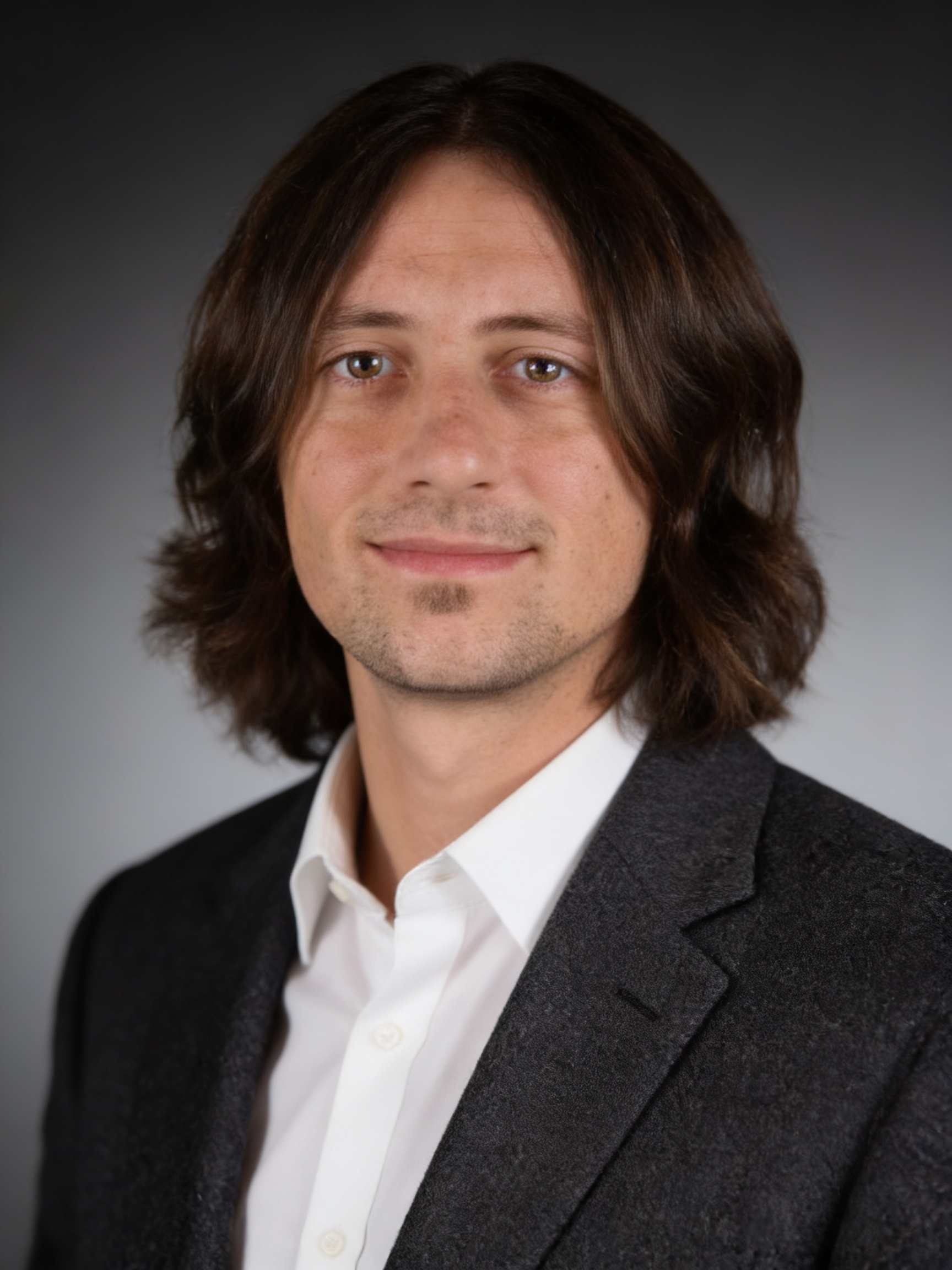 Headshot of a man with shoulder-length dark brown hair, light facial hair, wearing a black blazer over a white shirt, in front of a dark gray background.