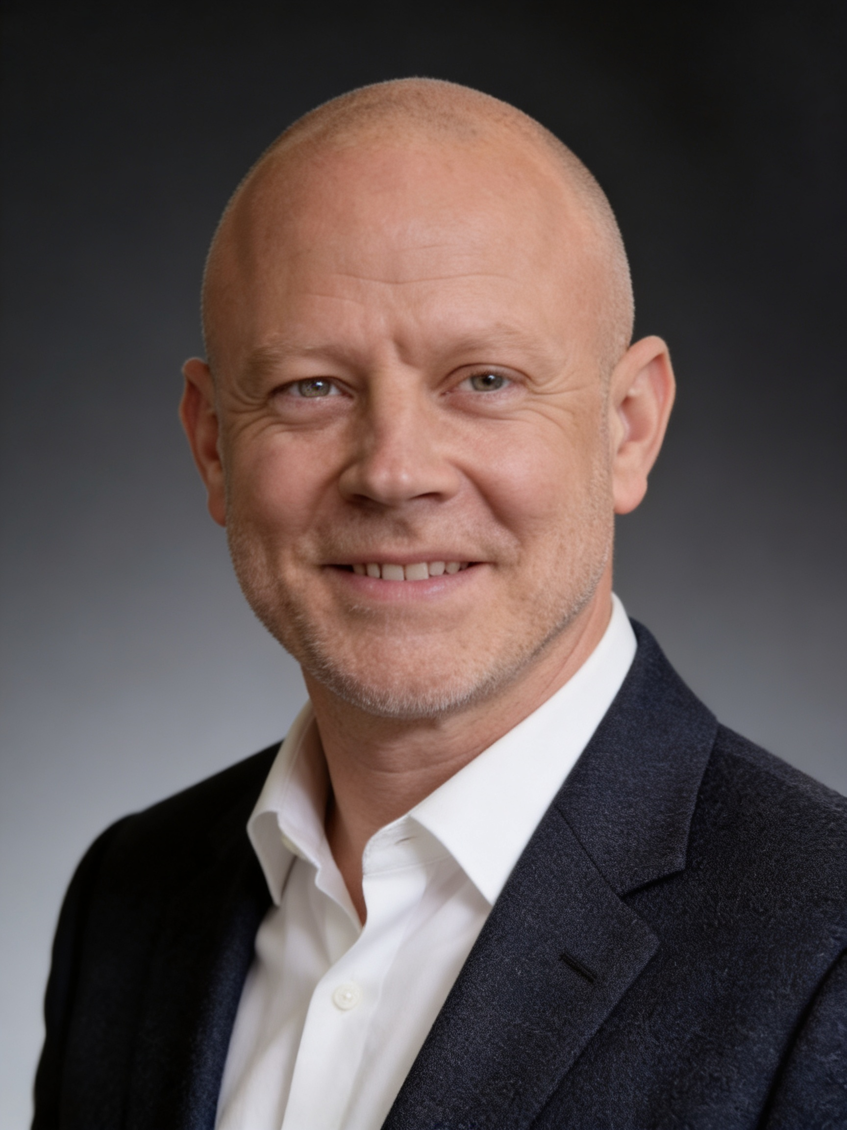 Close-up of a smiling bald man in a white shirt and dark blazer against a gray gradient background.
