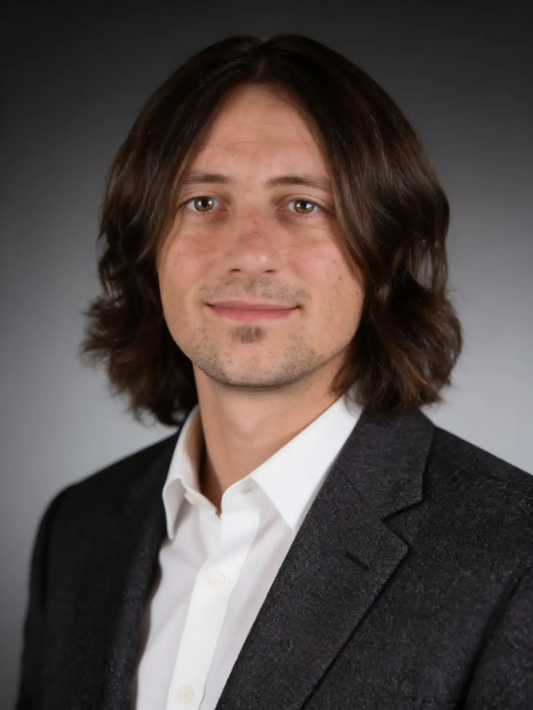 Headshot of a man with shoulder-length dark brown hair, light facial hair, wearing a black blazer over a white shirt, in front of a dark gray background.