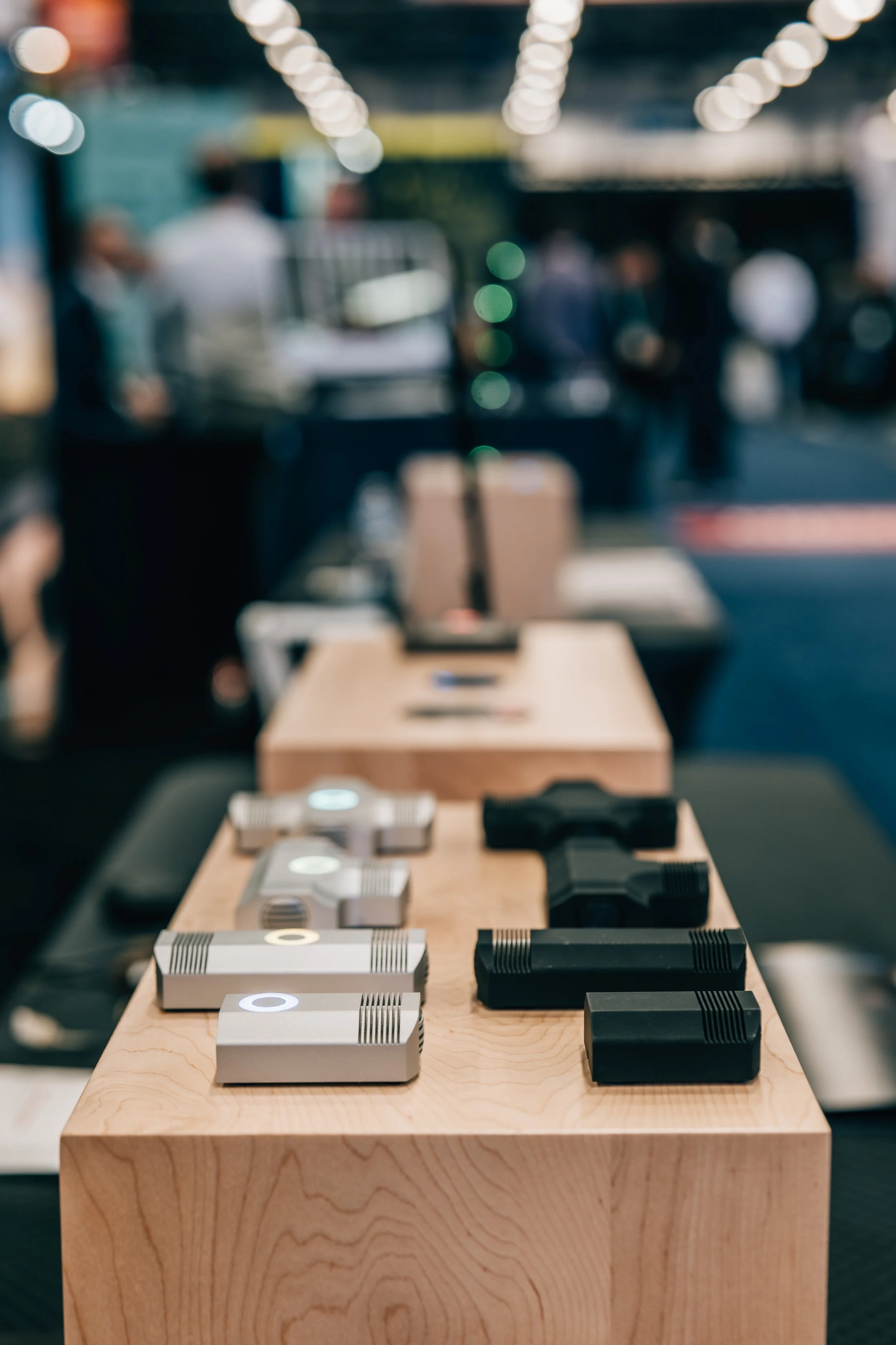 Display of various sleek, modern gadgets arranged on a wooden table, with blurred background of people and bright ceiling lights.