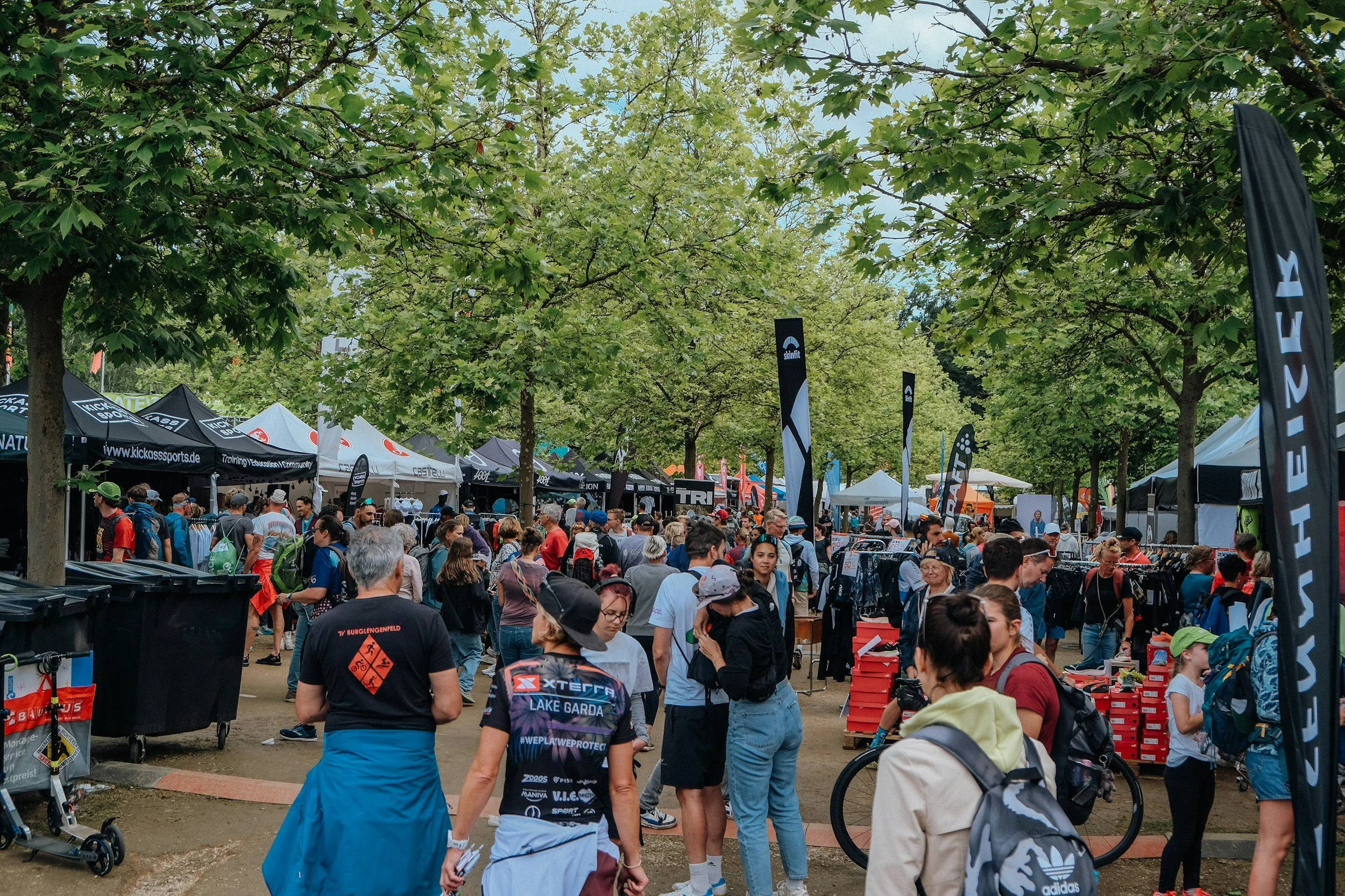 A crowded outdoor market or fair with numerous people walking and shopping under a canopy of green trees. Various vendor tents and flags are visible, suggesting a lively event.