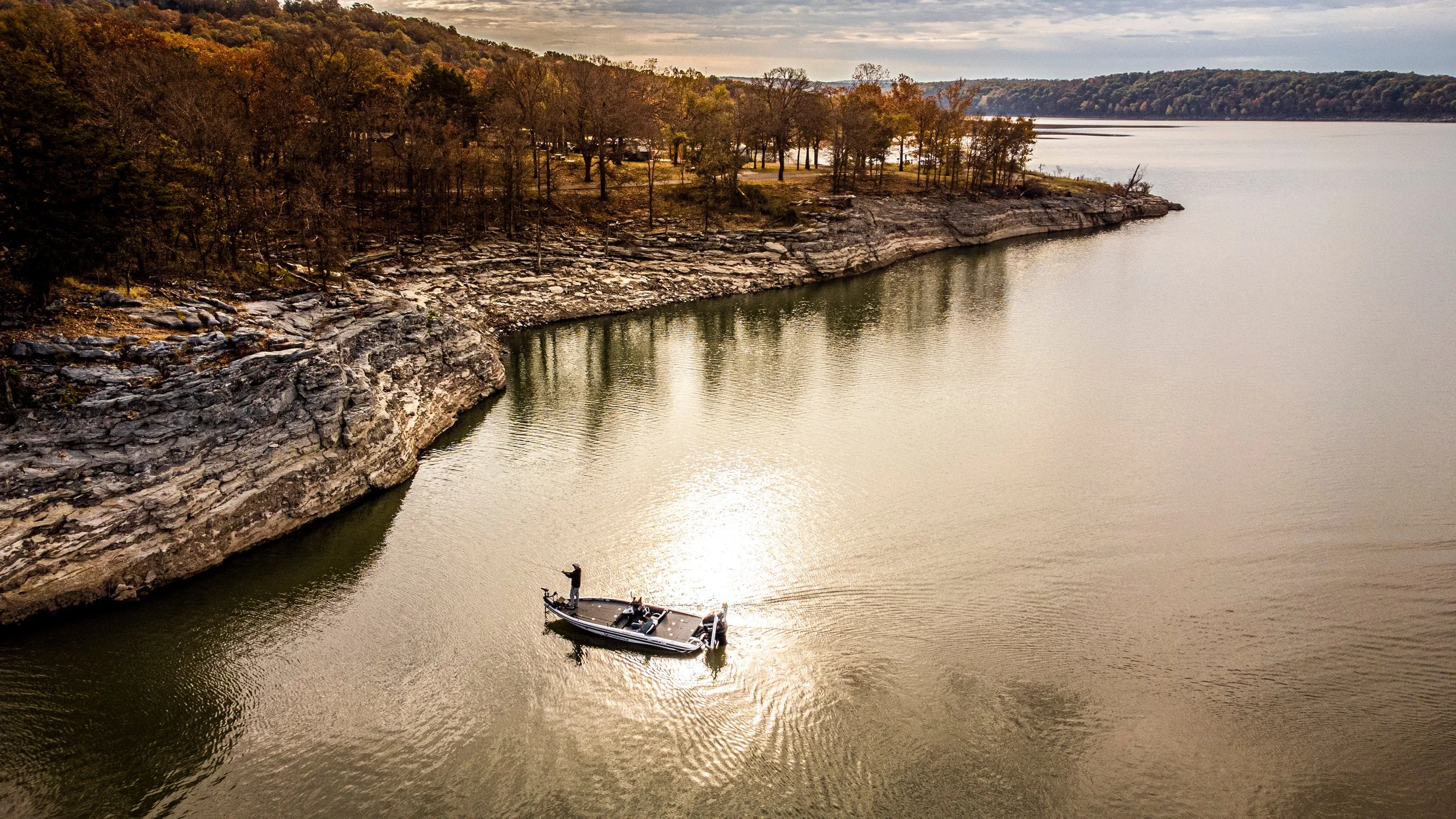 A small boat with a person fishing on a river with rocky shoreline and trees showing fall foliage in the background.