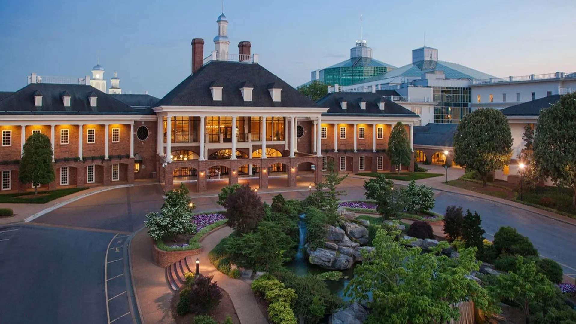 A large, elegant brick building with a classical architectural style, with multiple windows and a prominent entrance with tall white columns. The building is surrounded by landscaped gardens, trees, and a small waterfall feature, under a partly clear sky in the early evening with lighting illuminating the structure.