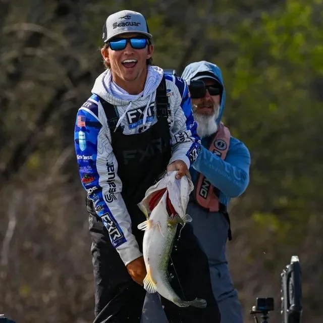 A man wearing a fishing vest, sunglasses, and a baseball cap holding a large fish with a big smile, standing outdoors with trees in the background.