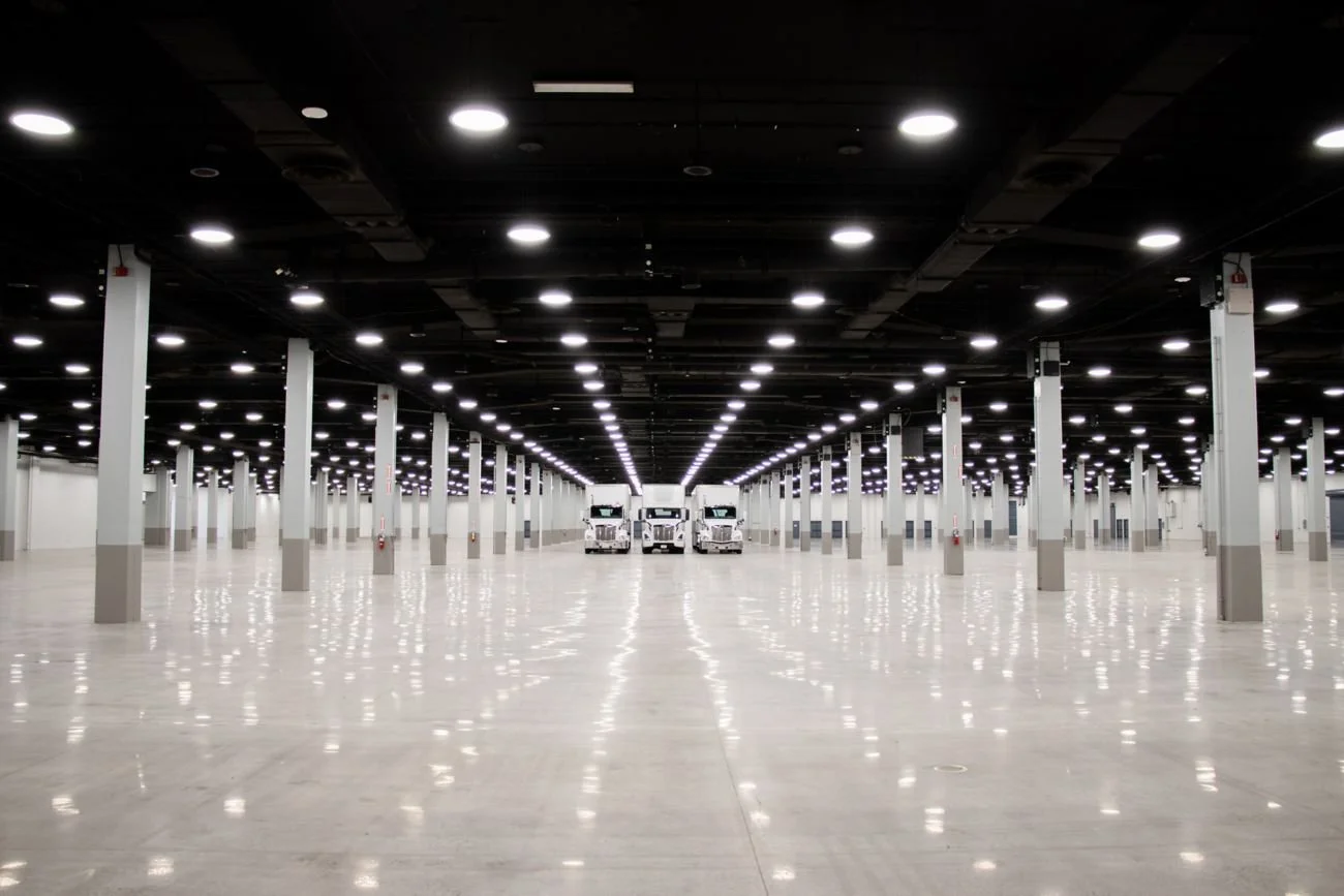 Empty large indoor warehouse or convention hall with bright overhead lights and three trucks in the distance.