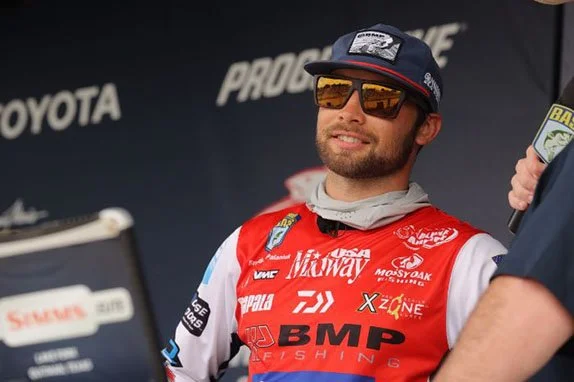A man wearing sunglasses, a cap, and a red racing suit with various logos, smiling during a racing event.