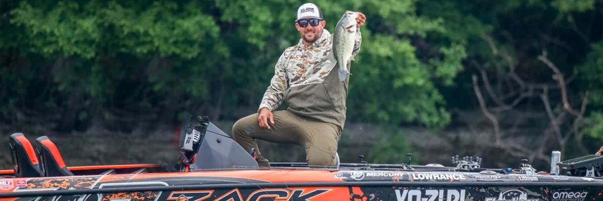 A man sitting in a boat holding up a largemouth bass fish he caught, with lush green trees in the background.
