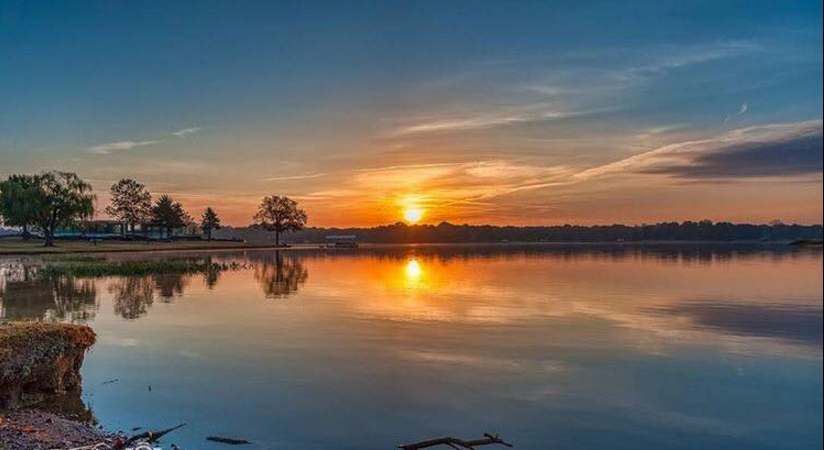 Sunset over a calm lake with trees on the shore and reflections in the water.
