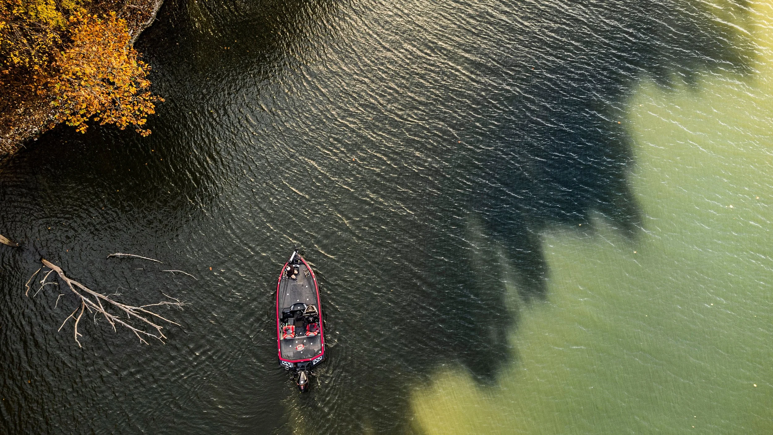A boat with two people on board navigating a body of water with contrasting dark and light areas, possibly indicating different water conditions or substances. A fallen tree branch is partially submerged along the shoreline on the left side of the image.
