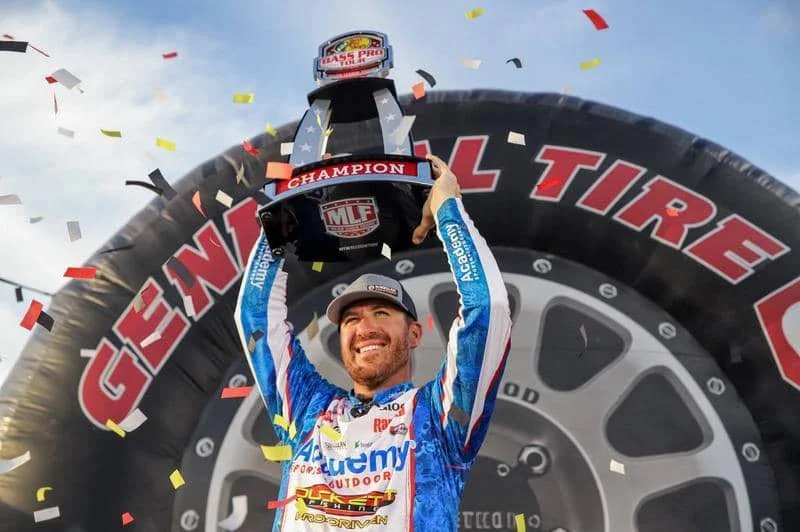 A race car driver in a blue and white racing suit holding up a trophy on the winner's podium, with a large tire and colorful confetti in the background.