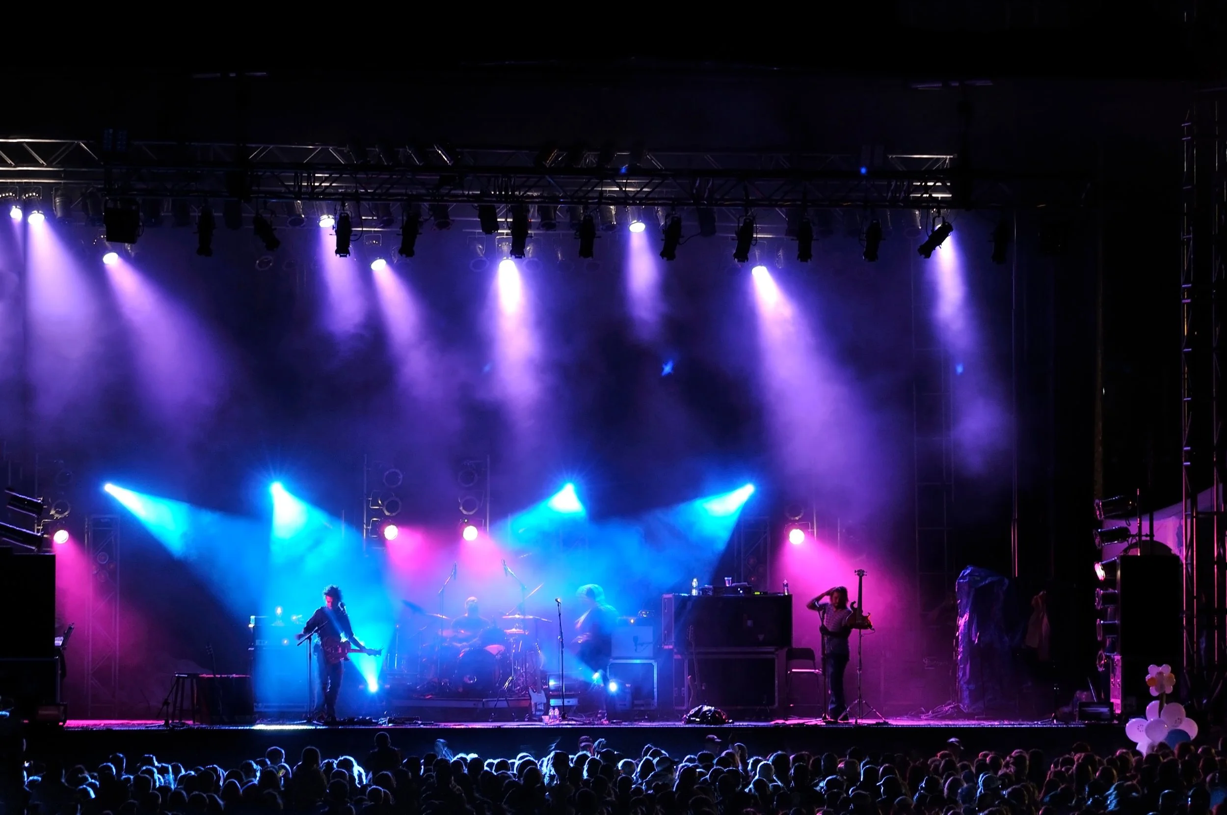 Stage with musicians performing under purple and blue lights, silhouetted audience in foreground.