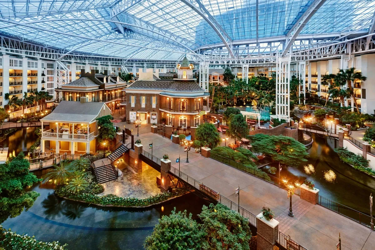 Indoor shopping mall with a glass ceiling, featuring a water canal with small bridges, surrounded by shops, trees, and seating areas, illuminated with warm lighting.