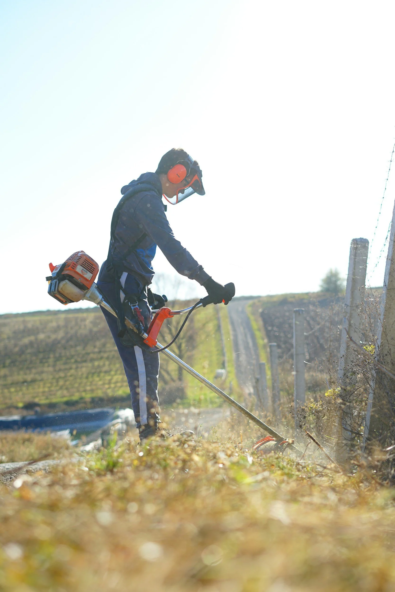 Person wearing safety goggles, ear protection, and a backpack trimmer cutting grass near a fence post on a hillside.