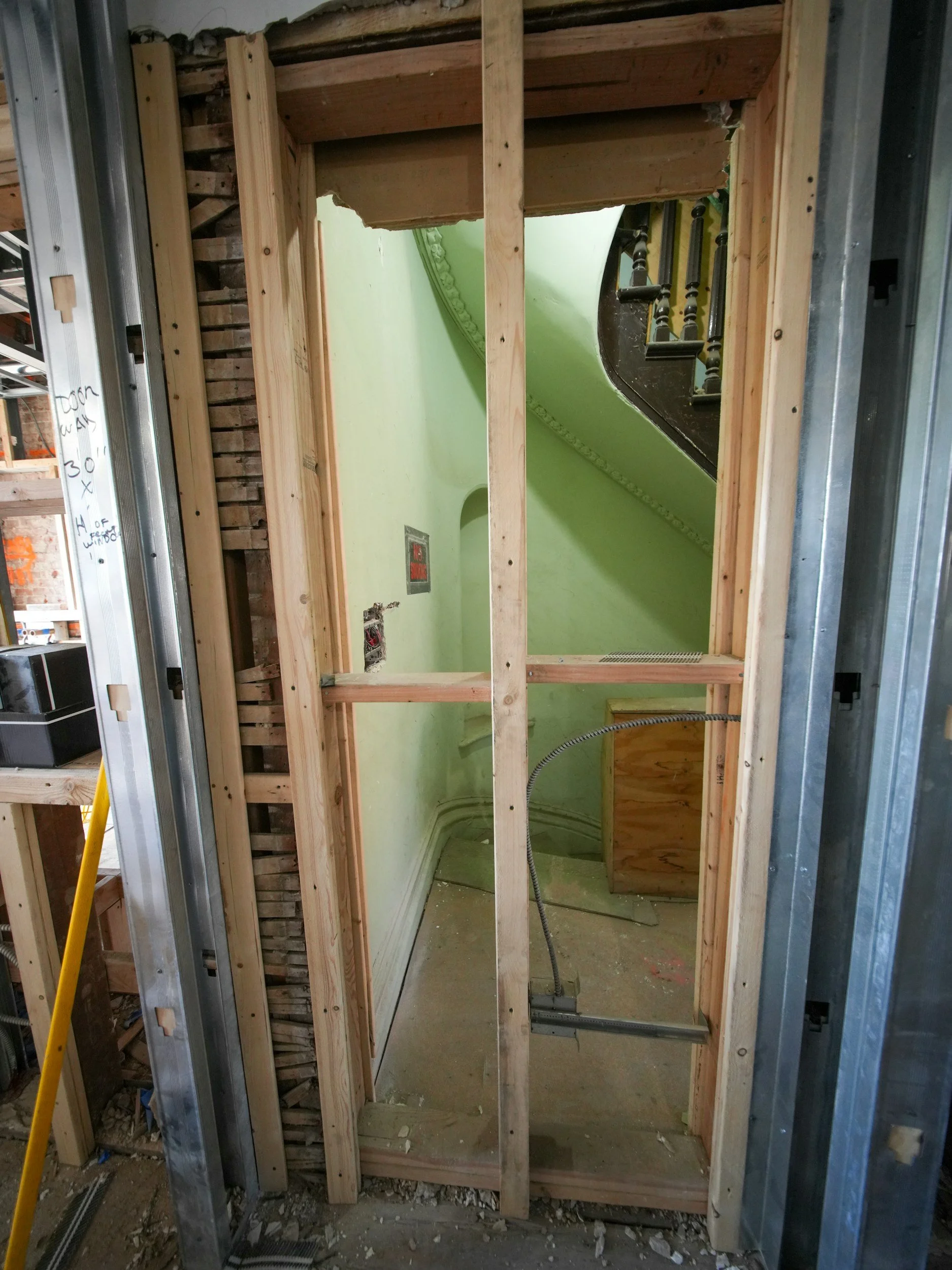 Interior view of a partially built wall with wooden framing and a staircase with dark wooden banister and spindles in the background. The floor is unfinished concrete with construction debris.