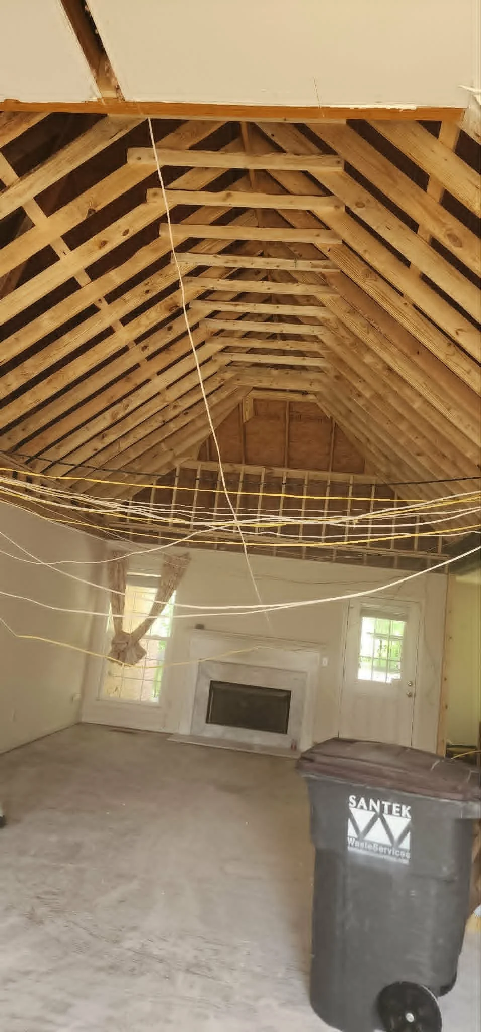 Interior of a room under renovation with exposed wooden rafters and beams in the ceiling, hanging strings or wires, a window, a fireplace, and a black trash bin labeled 'SANTEK Water Services'.