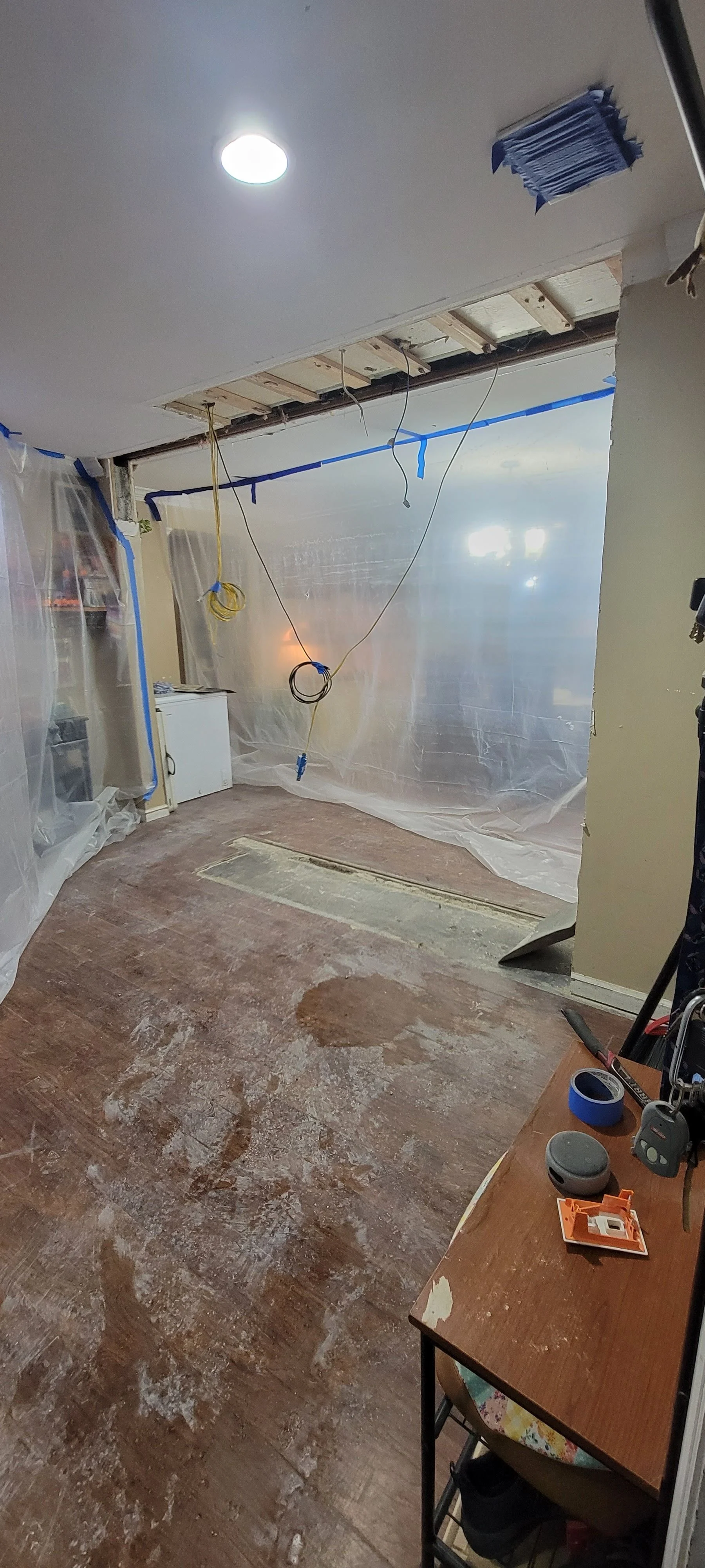 Room under renovation with exposed ceiling, plastic sheeting, wires hanging, and construction tools and debris on the floor.