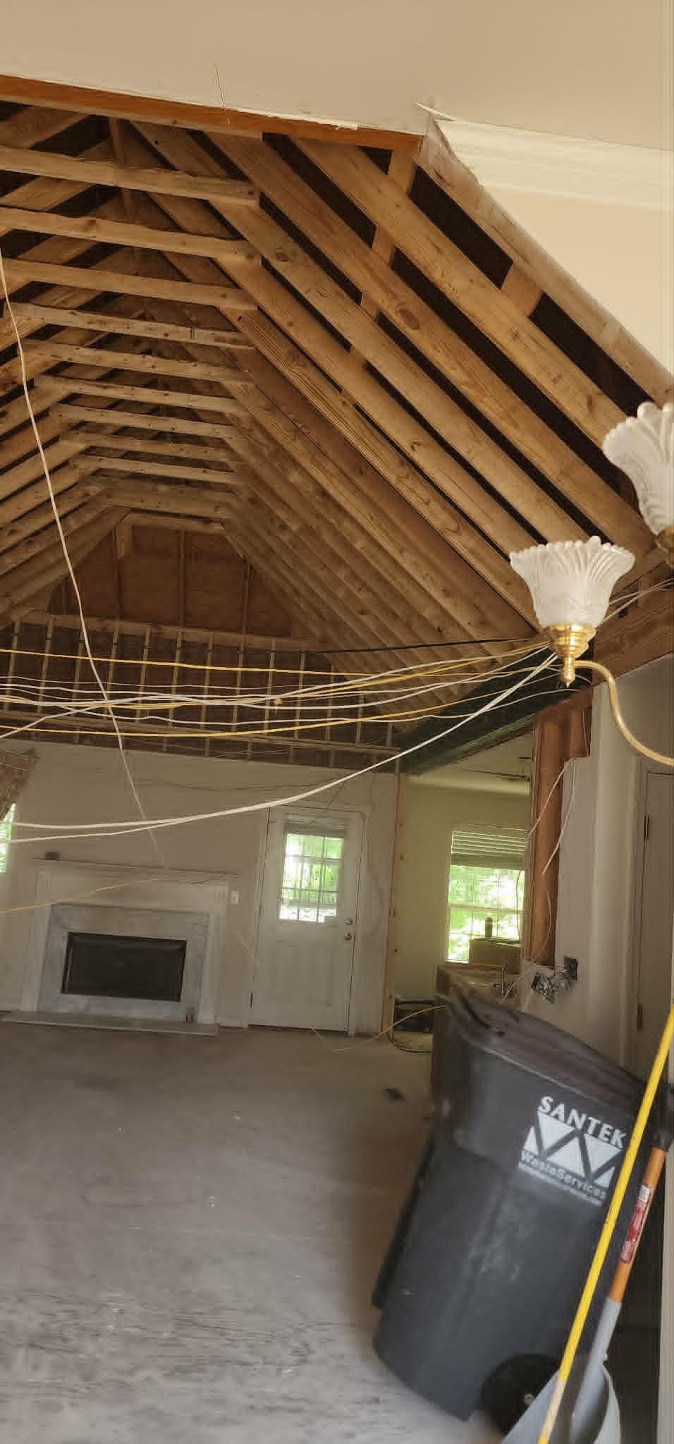 Interior view of an unfinished room with exposed ceiling framing, a hanging light fixture, a trash can, and construction wiring.