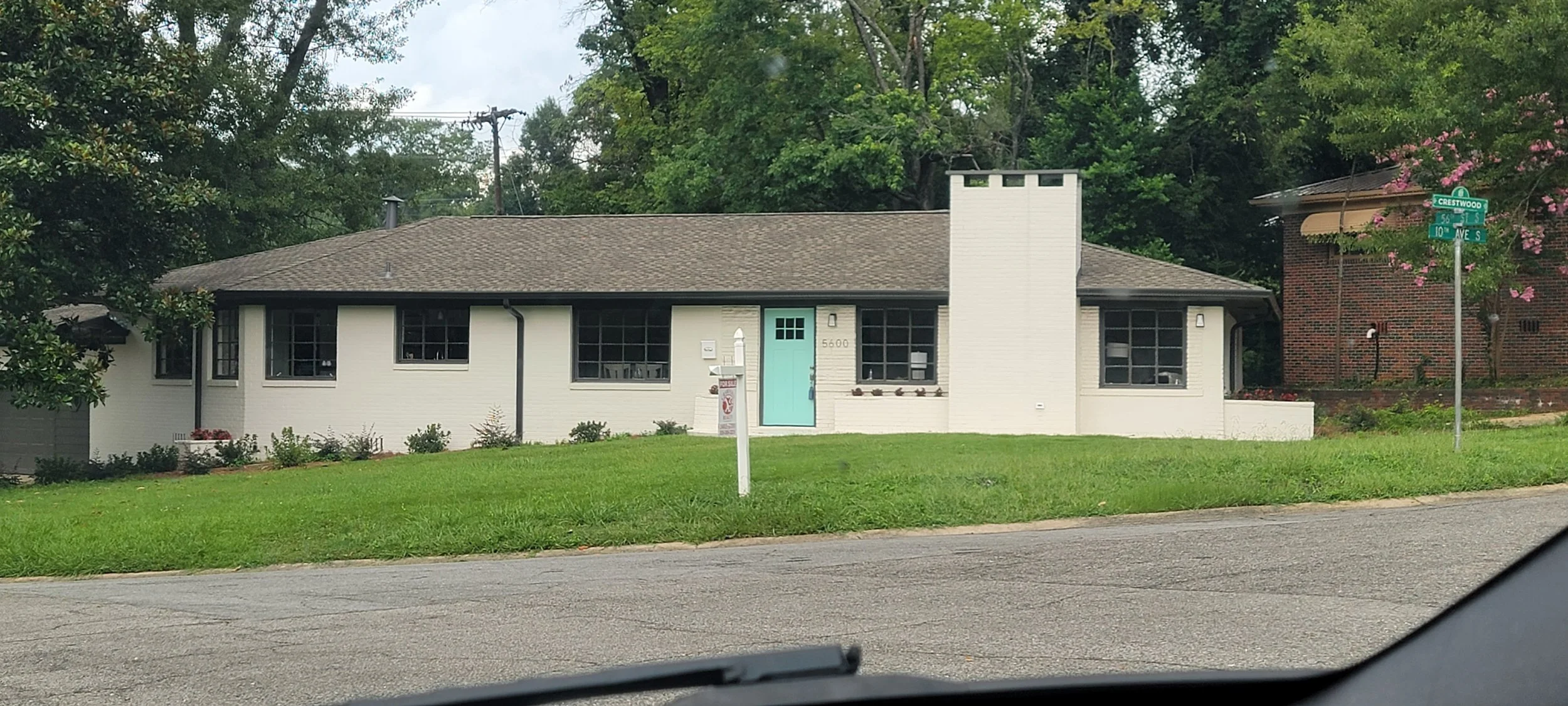 A single-story white house with black window frames and a turquoise front door, surrounded by green grass and trees, on a corner with street signs for Crestwood and 10th Avenue South.