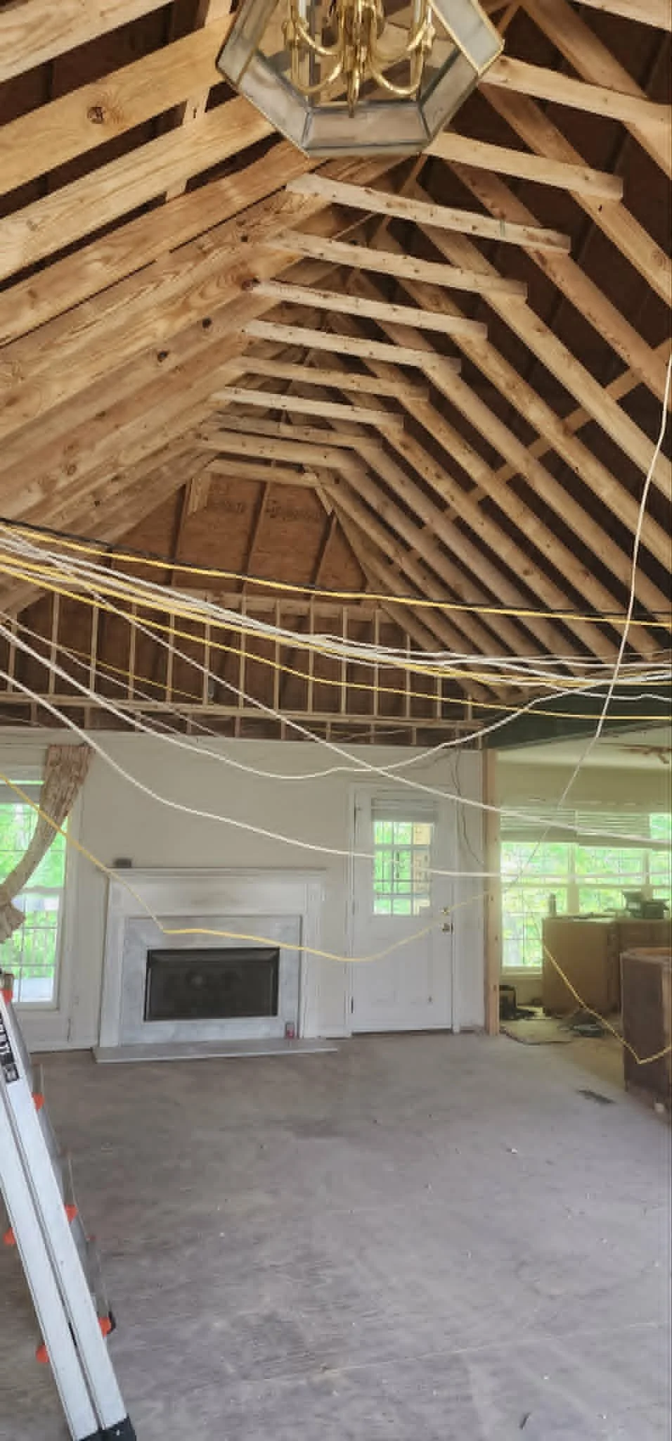 Interior of a house under renovation with exposed wooden roof rafters, electrical wires hanging, a white fireplace, and unfinished walls.