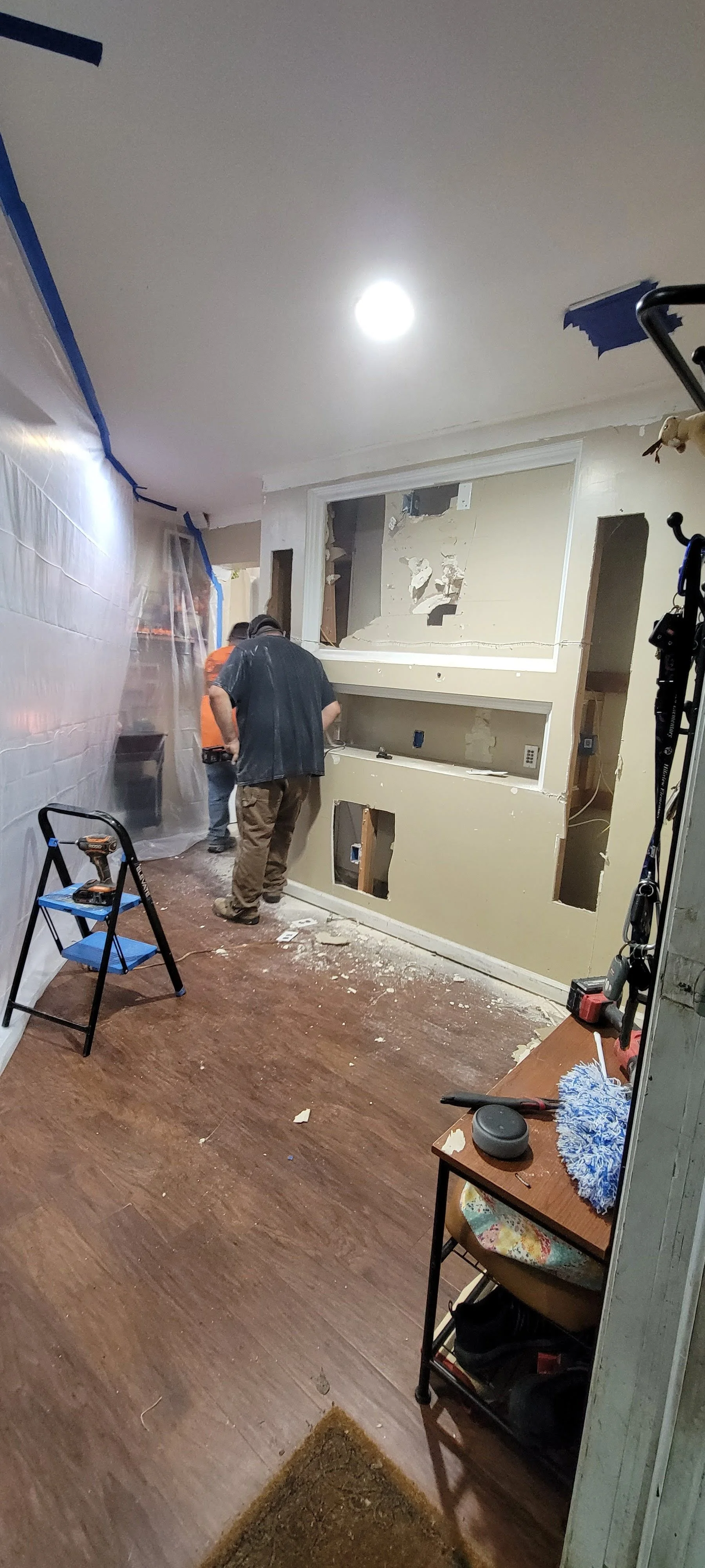 Construction workers repairing a wall inside a house, with tools and debris on the floor, plastic sheeting covering part of the area, and renovation equipment present.