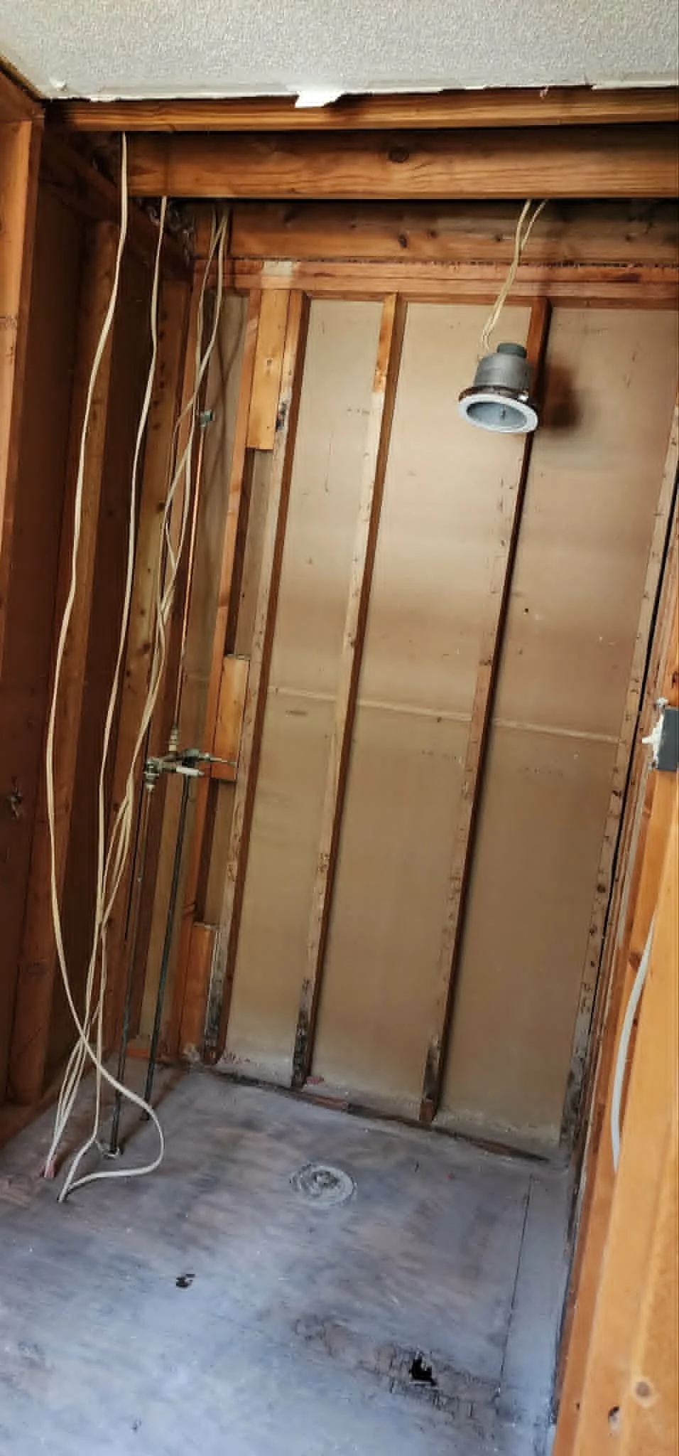 Renovation of a small room with exposed wooden wall studs, electrical wires hanging loosely, and a ceiling fixture without a cover. The floor is concrete with some debris and a drain in the center.