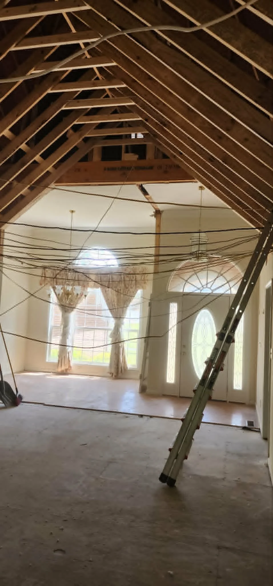 Interior of a house under construction with exposed wooden roof framing, hanging electrical wires, a ladder, and a large window with curtains.