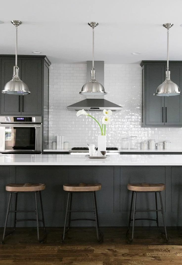 A modern kitchen featuring a white countertop island with three wooden and metal stools, dark gray cabinets, a white tiled backsplash, stainless steel oven and microwave, a stainless steel range hood, and three metallic pendant lights hanging from th