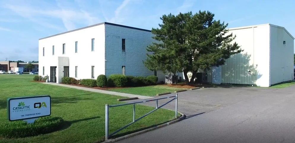Industrial building with a sign for "Catalytic Generators, QA" located on Pineridge Road, lawn and trees in the foreground, clear sky above.