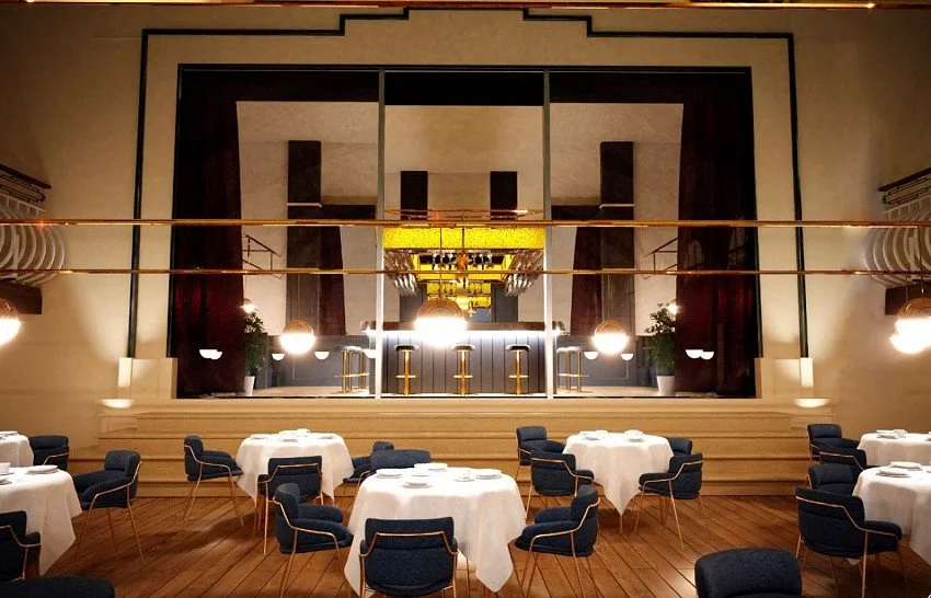 Elegant restaurant dining area with round tables covered in white tablecloths, surrounded by blue chairs with wooden legs, large decorative lighting fixtures, and a raised stage with curtains behind it.