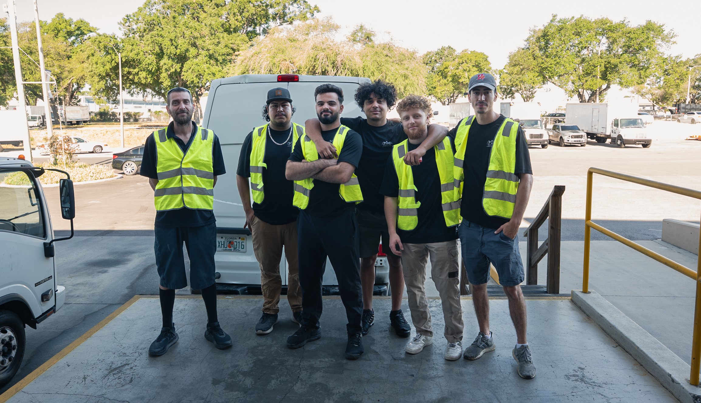 Group of six young men standing together outdoors, some wearing neon safety vests, in front of a white van, with trees and parked cars in the background.
