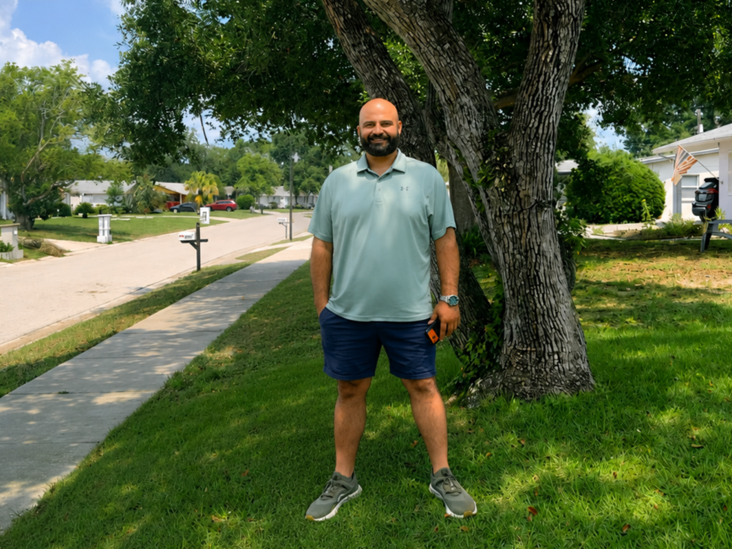 Man standing on grass in front of tree on suburban street, smiling, holding a phone, wearing a light blue polo shirt, navy shorts, and sneakers.